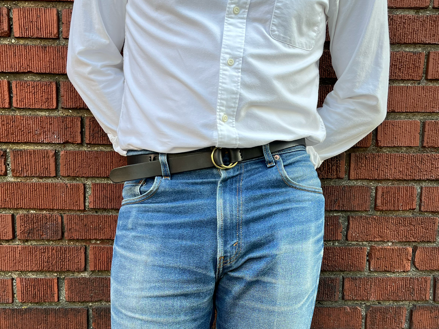 A person in a white button-down shirt and blue jeans stands against a red brick wall with hands behind their back. The shirt is untucked and they are wearing a black belt.