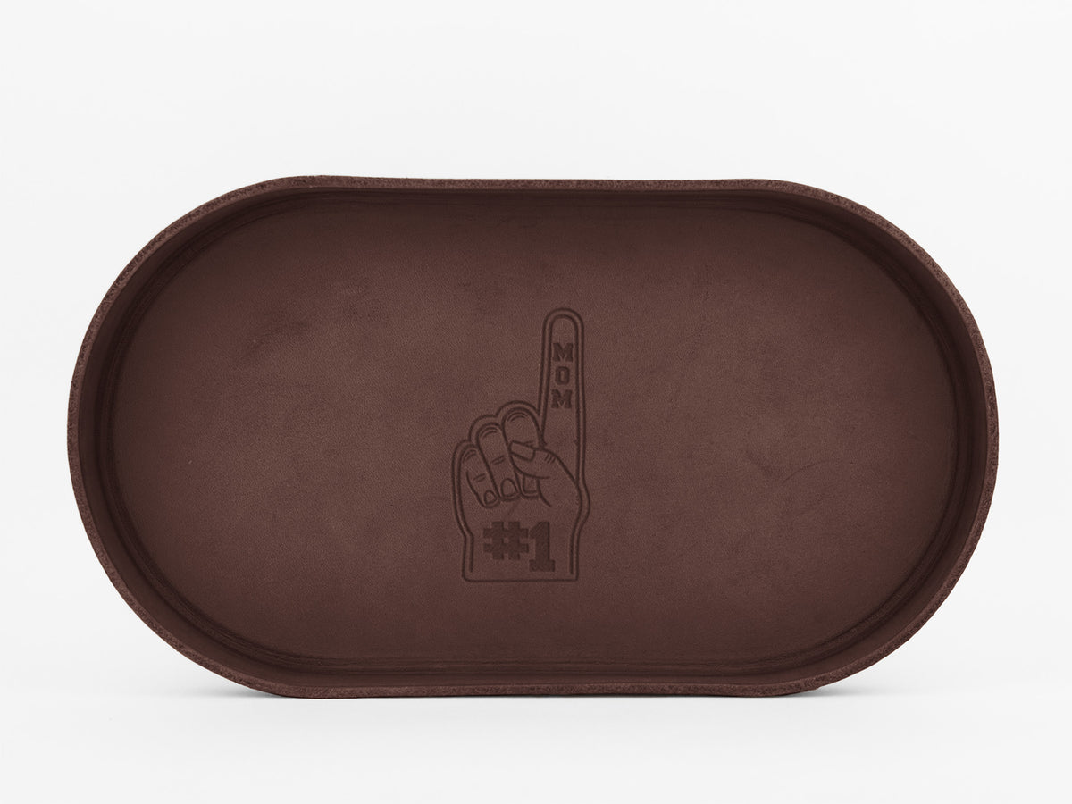 A brown, oval-shaped tray with an embossed design of a foam finger. The finger points upward and has the words “#1 MOM” written on it. The tray is empty and placed against a plain white background.