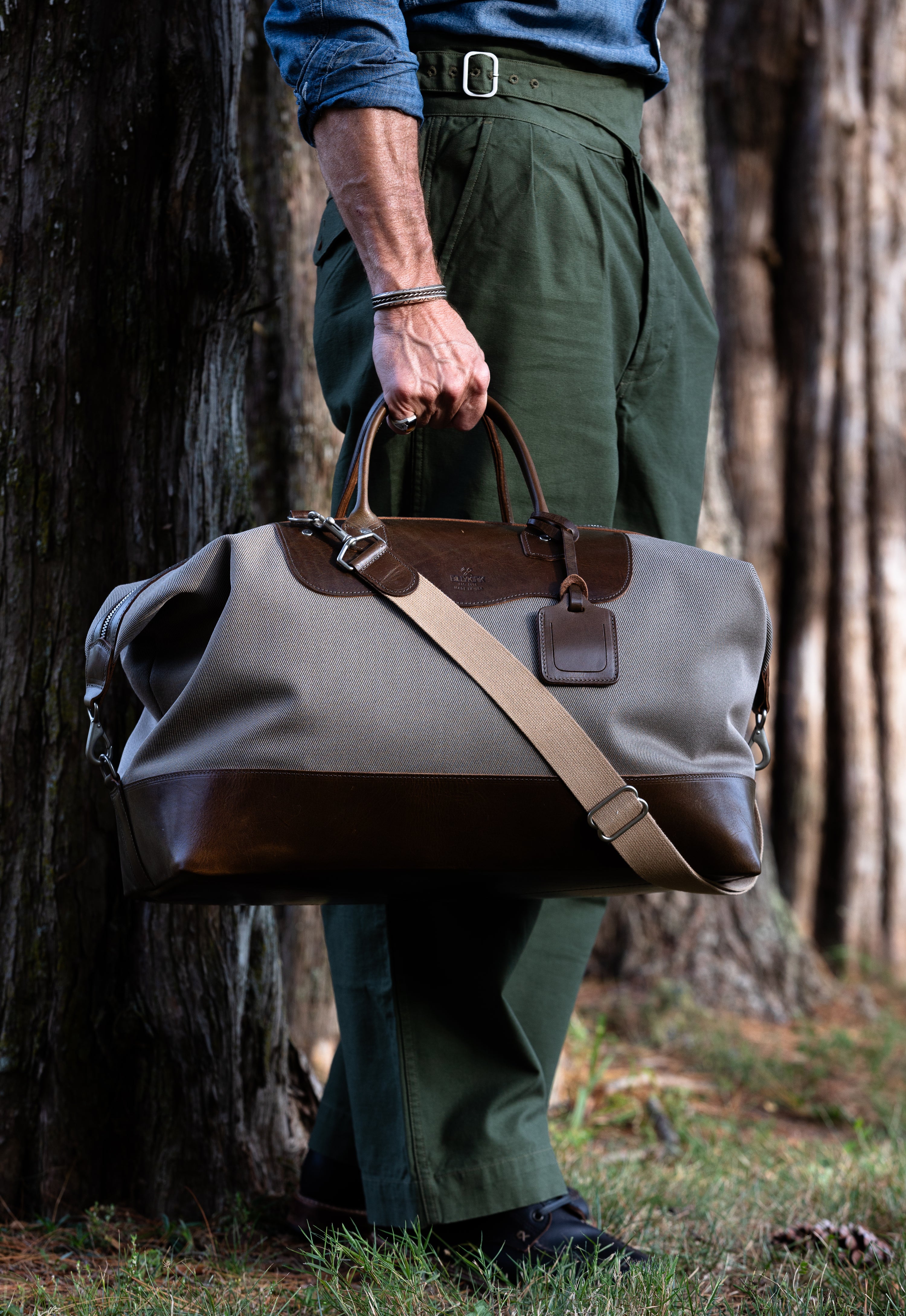 Person holding a beige duffel bag with olive leather accents in a forest setting