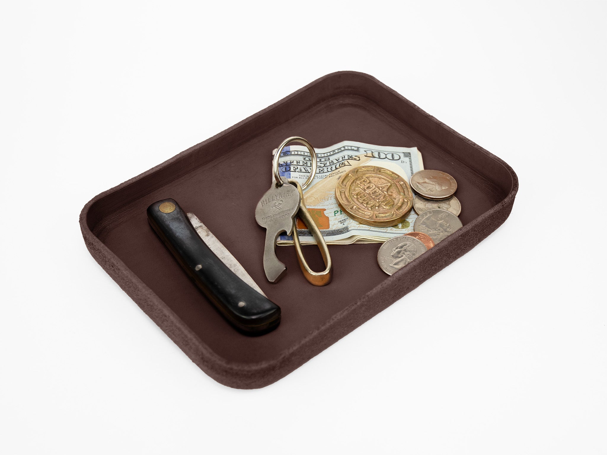 A brown tray holding a straight razor, a folding bottle opener, a $100 bill, coins, and a brass key on a white background.