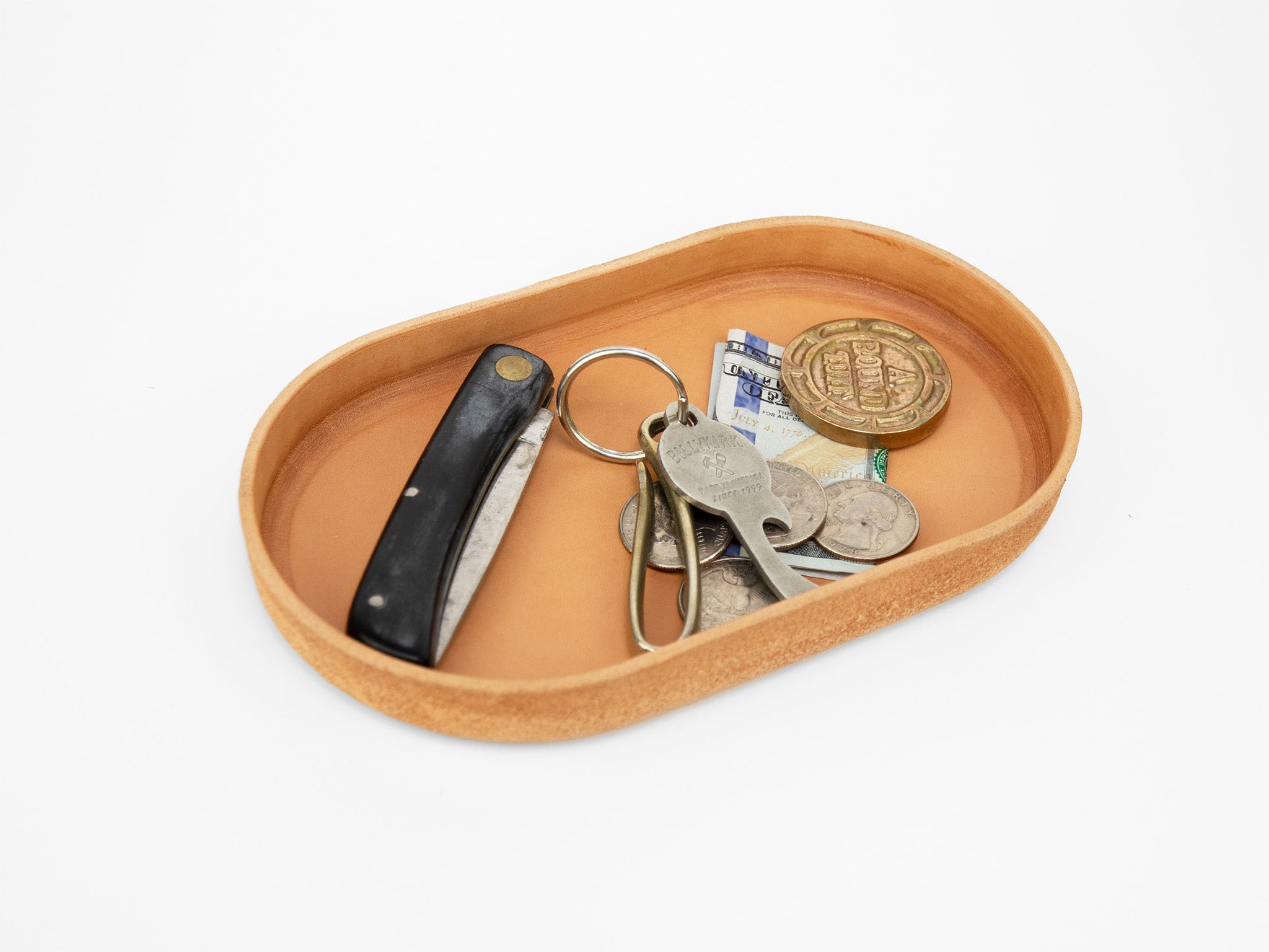 A tan leather tray holding a folding pocket knife, a keychain with keys, assorted coins, a folded banknote, and a large round token, all placed on a white background.
