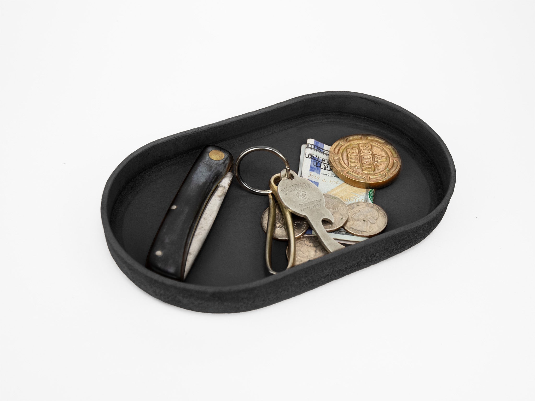 A black tray containing a folding pocket knife, a set of keys, coins, a folded banknote, and a large gold token, all set against a white background.