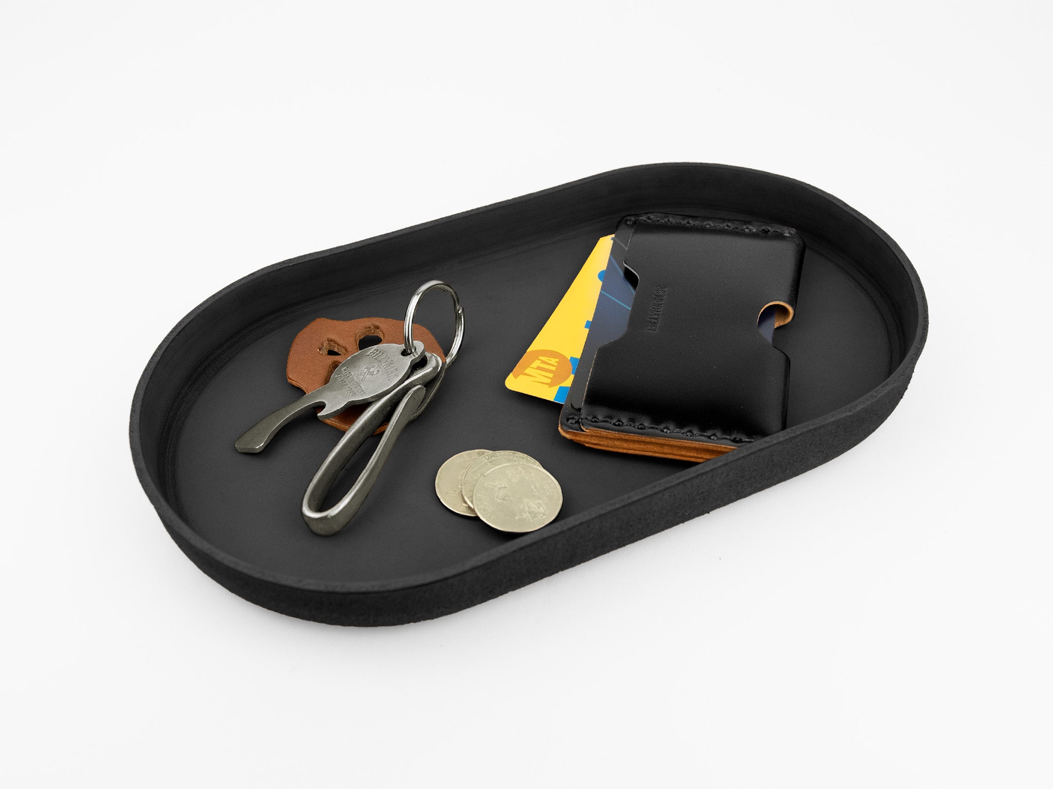 A black oval tray holding a leather card holder with visible cards, several coins, a set of keys with a leather keychain, and a metal key clip, all on a white background.