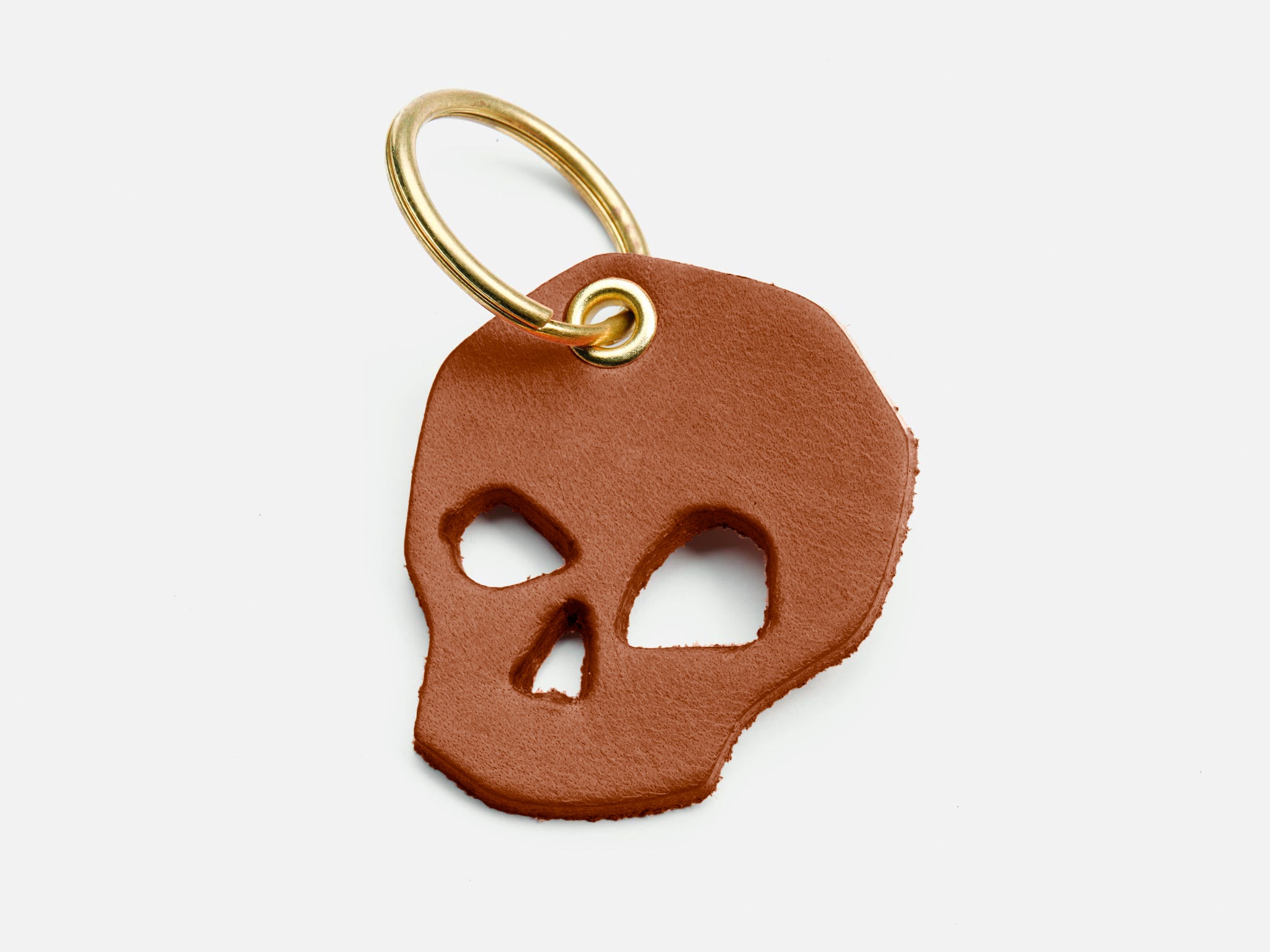 A keychain featuring a brown leather charm cut into the shape of a skull with hollowed eyes and nose, attached to a gold metal ring, on a white background.