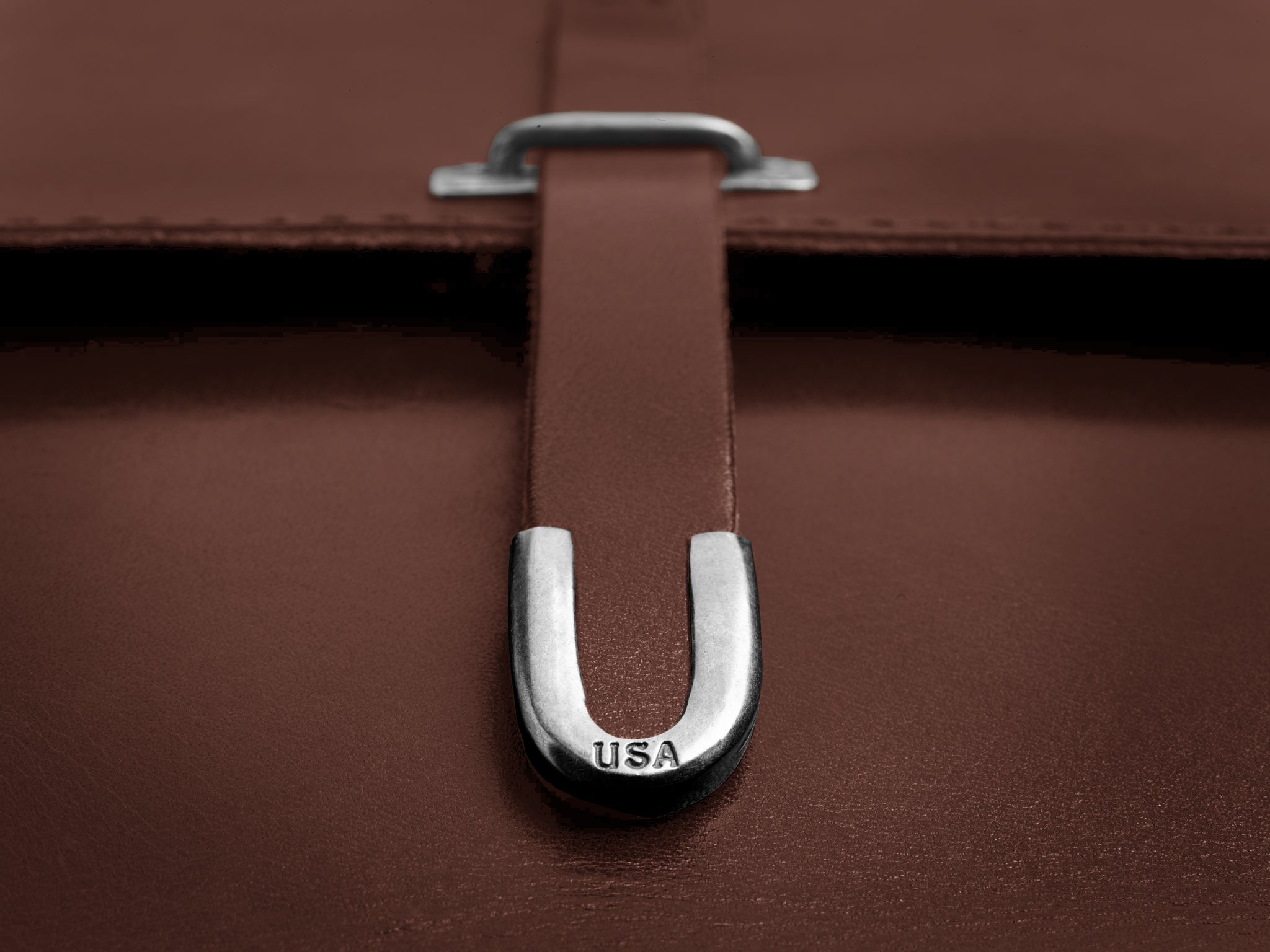 Close-up of a brown leather bag with a metal U-shaped clasp engraved with the letters USA. The clasp secures a leather strap on the bag.