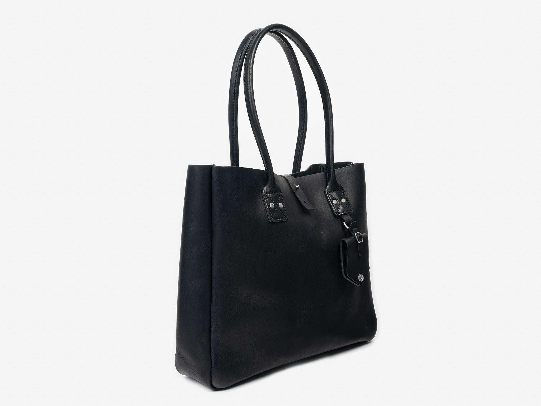 3/4 view of a medium-sized black leather tote with handles for your shoulder. The tote features a removable luggage tag below one of the handles. The bag is set against a neutral background.