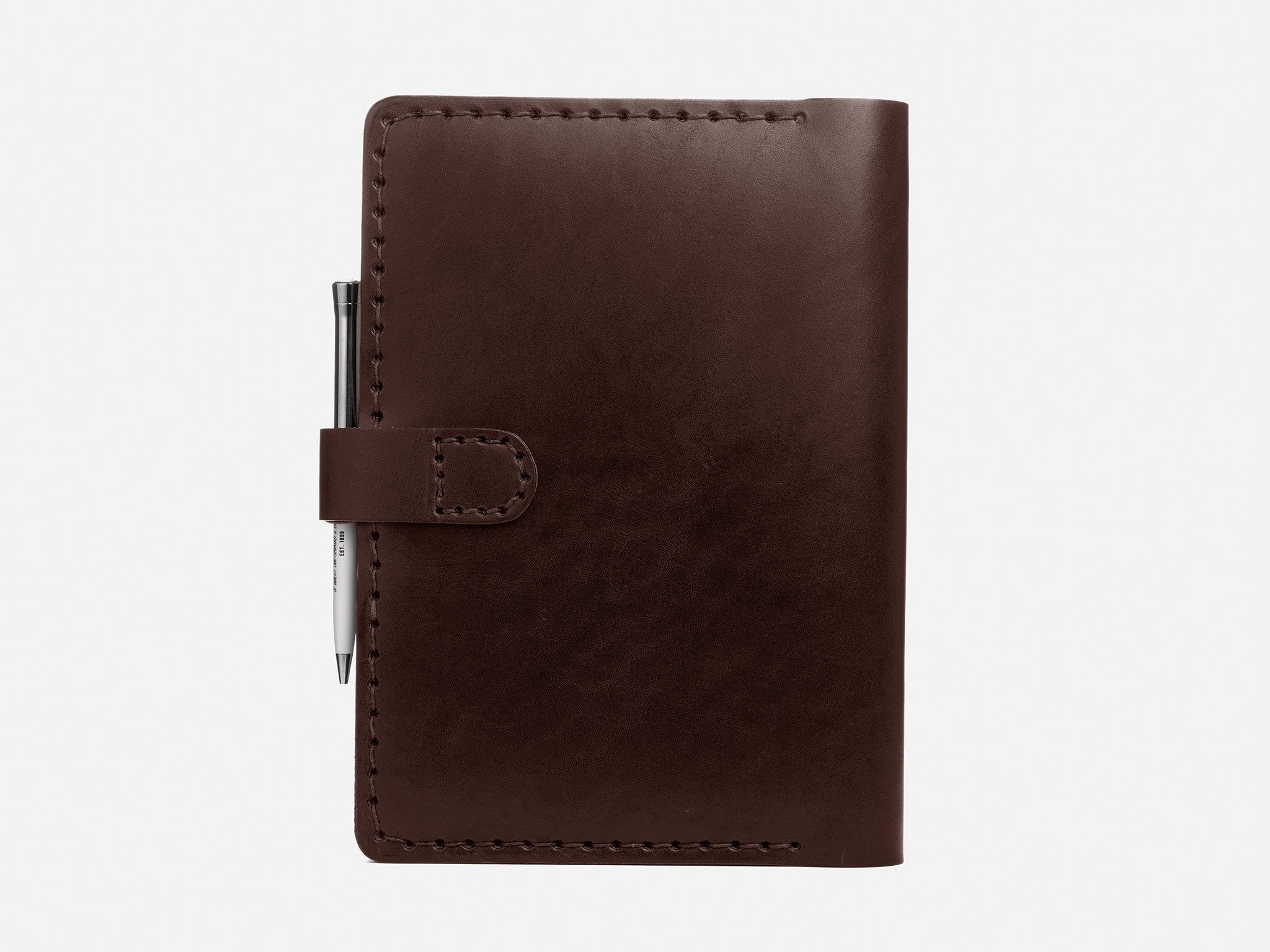A closed brown leather notebook with visible stitching, secured by a tab closure, with a silver pen attached on the left side, set against a plain white background.