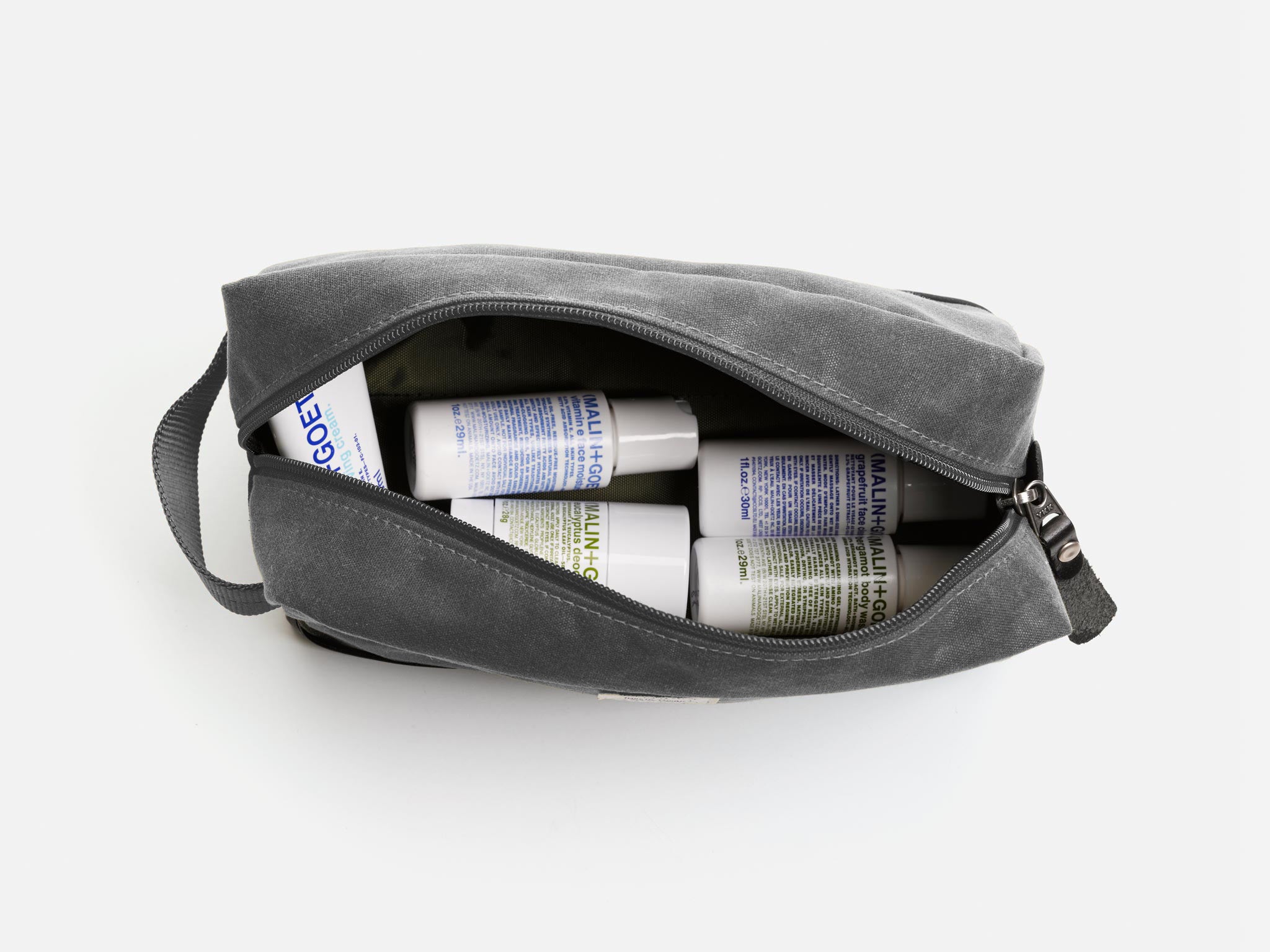 An ash waxed canvas toiletry bag unzipped to reveal several Malin+Goetz branded bottles and containers inside, arranged neatly against a white background.