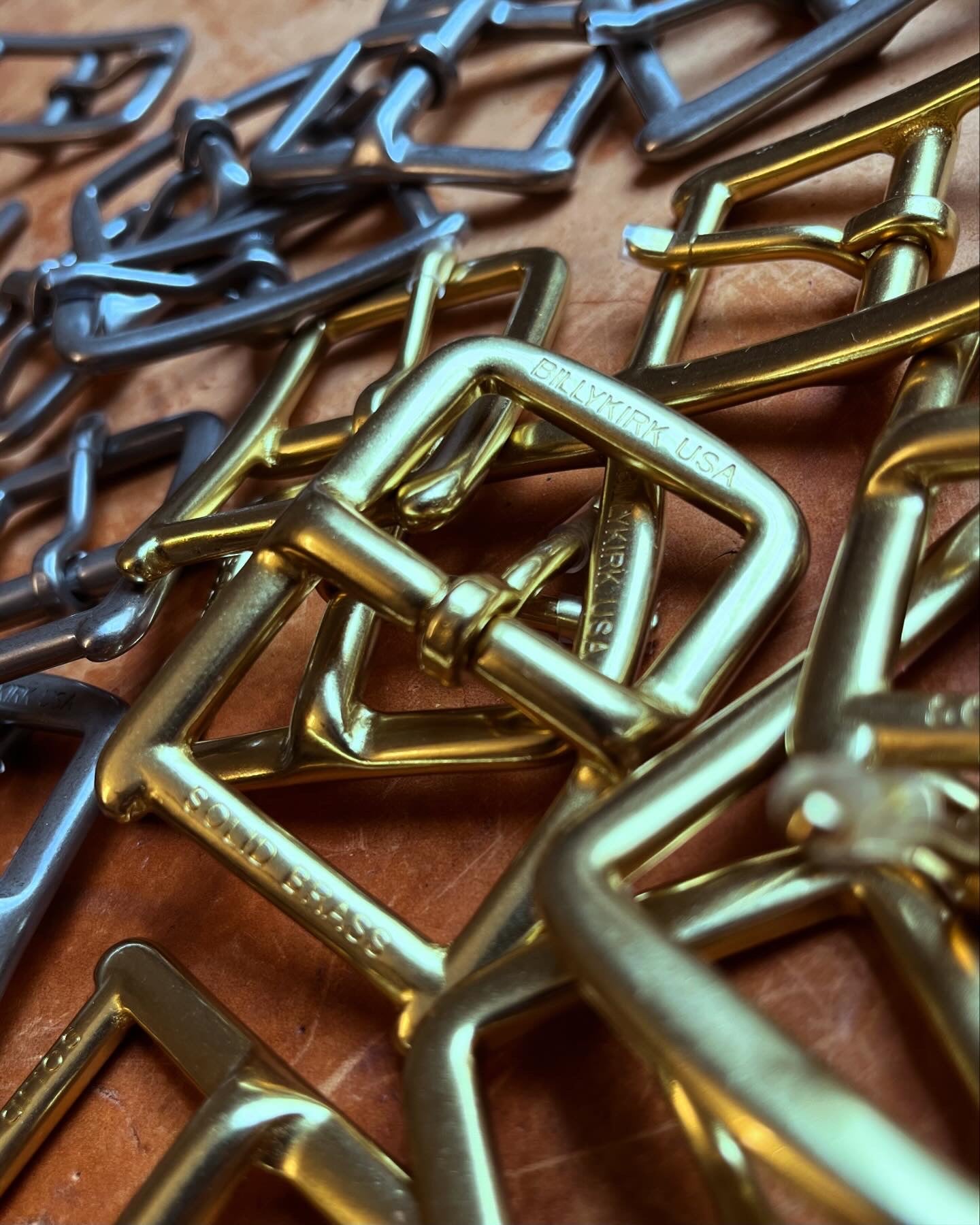 A close-up of several metal belt buckles, both gold and silver in color, arranged on a brown leather surface. Some buckles are engraved with “SOLID BRASS” and “BUCKLEMAN USA.”.