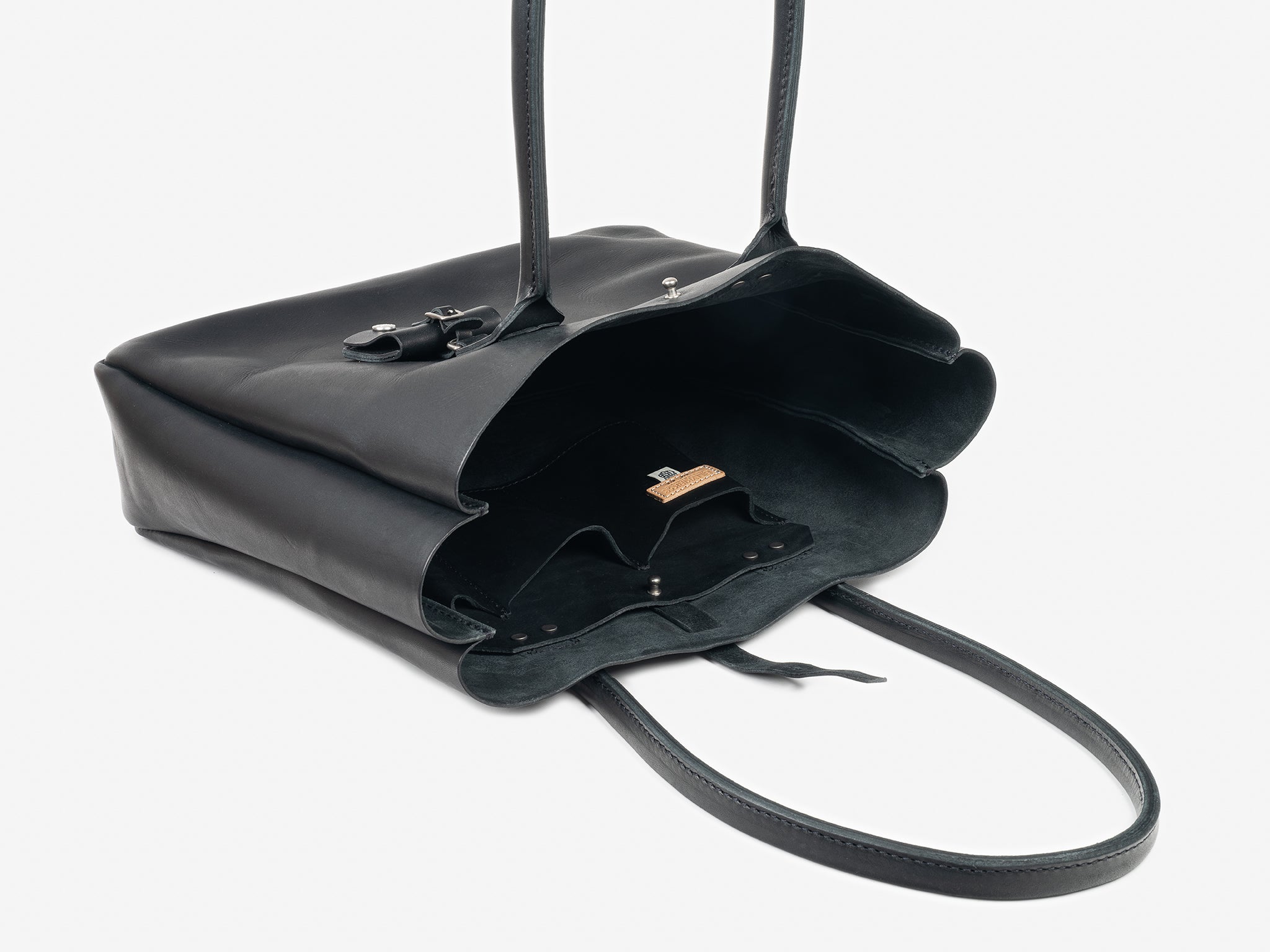 A black leather tote resting on its side, open. It features an interior organization pocket and a removable luggage tag set with a neutral background.