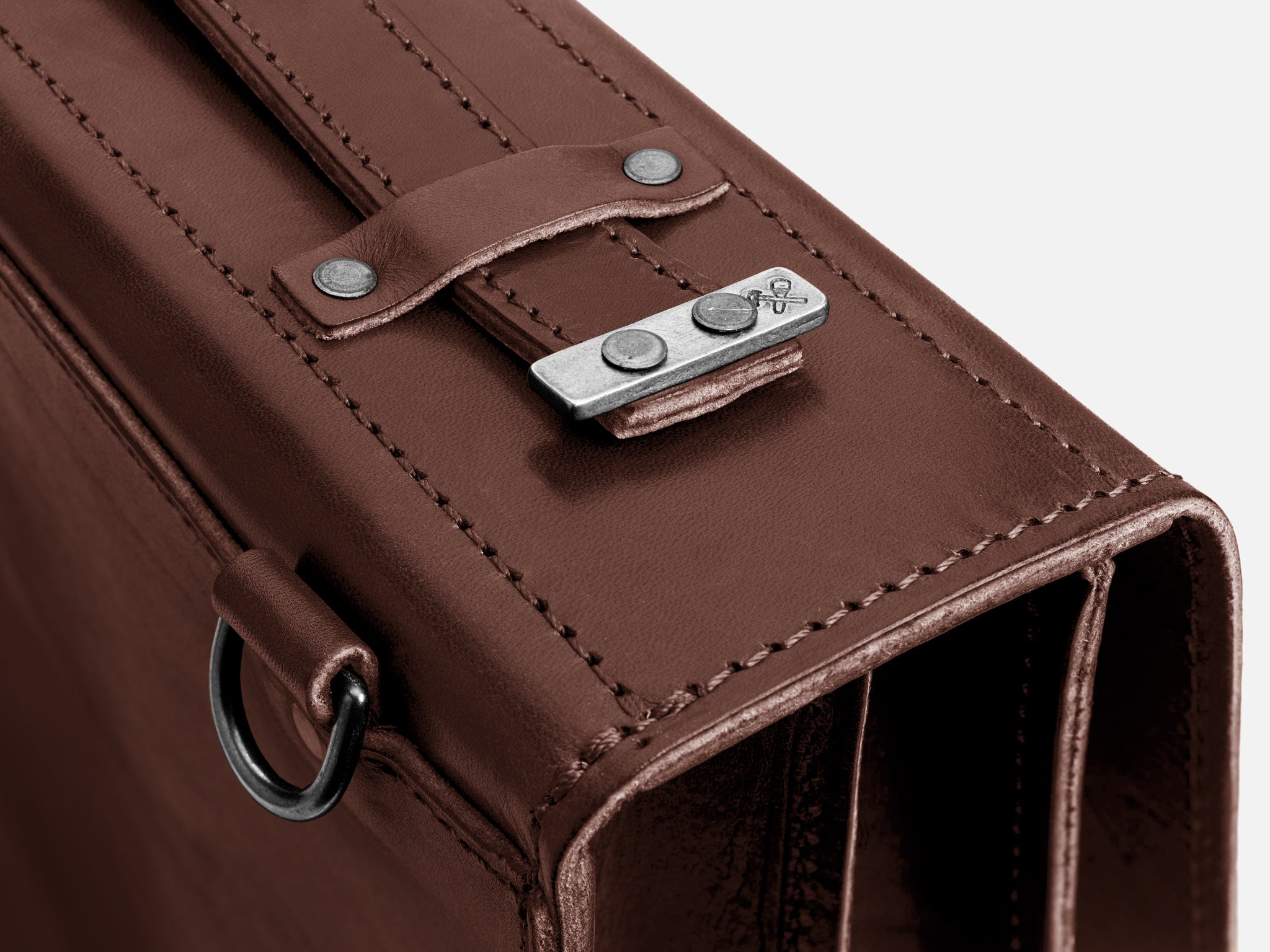 Close-up of a brown leather binder or briefcase with visible stitching, a metal clasp with two rivets, and a small embossed logo on the clasp. A metal D-ring is attached to the side.