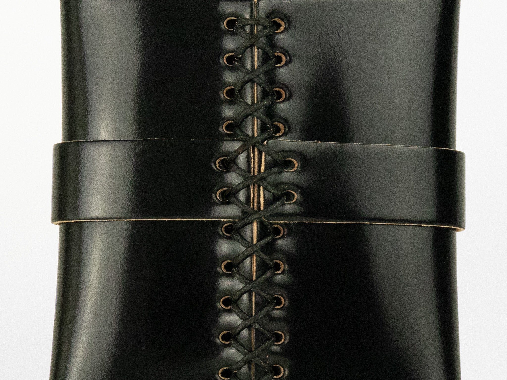Close-up of black leather boots featuring intricate crisscross lacing through metal eyelets, with a horizontal leather strap across the front.
