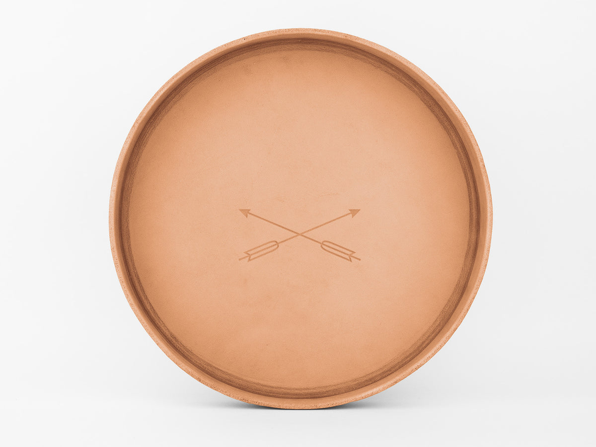 A round, tan clay plate with two crossed arrows engraved in the center, set against a plain white background.