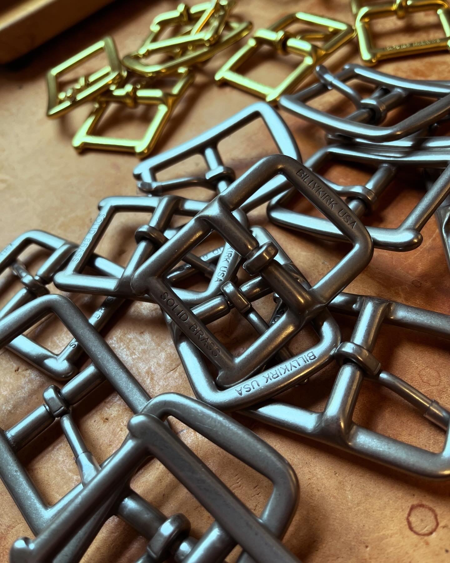 Close-up of overlapping silver and gold metal belt buckles on a tan surface. Some buckles have BILLYKIRK USA engraved on them. The scene is well-lit, highlighting the metallic textures.