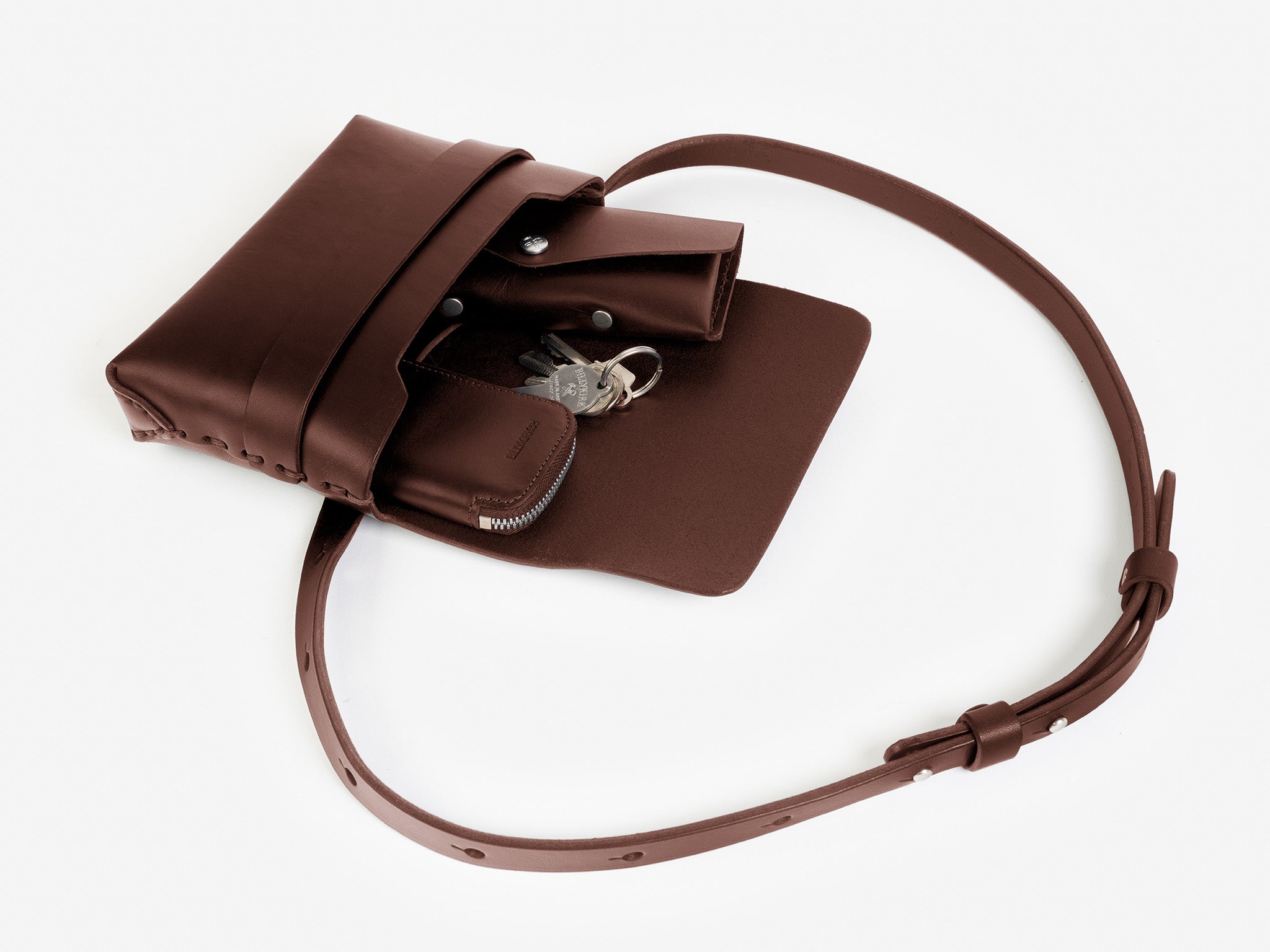 A hickory leather crossbody bag with a flap open, revealing a zippered coin pouch and a set of keys inside. The bag has a long adjustable strap and minimalist design, displayed on a white background.