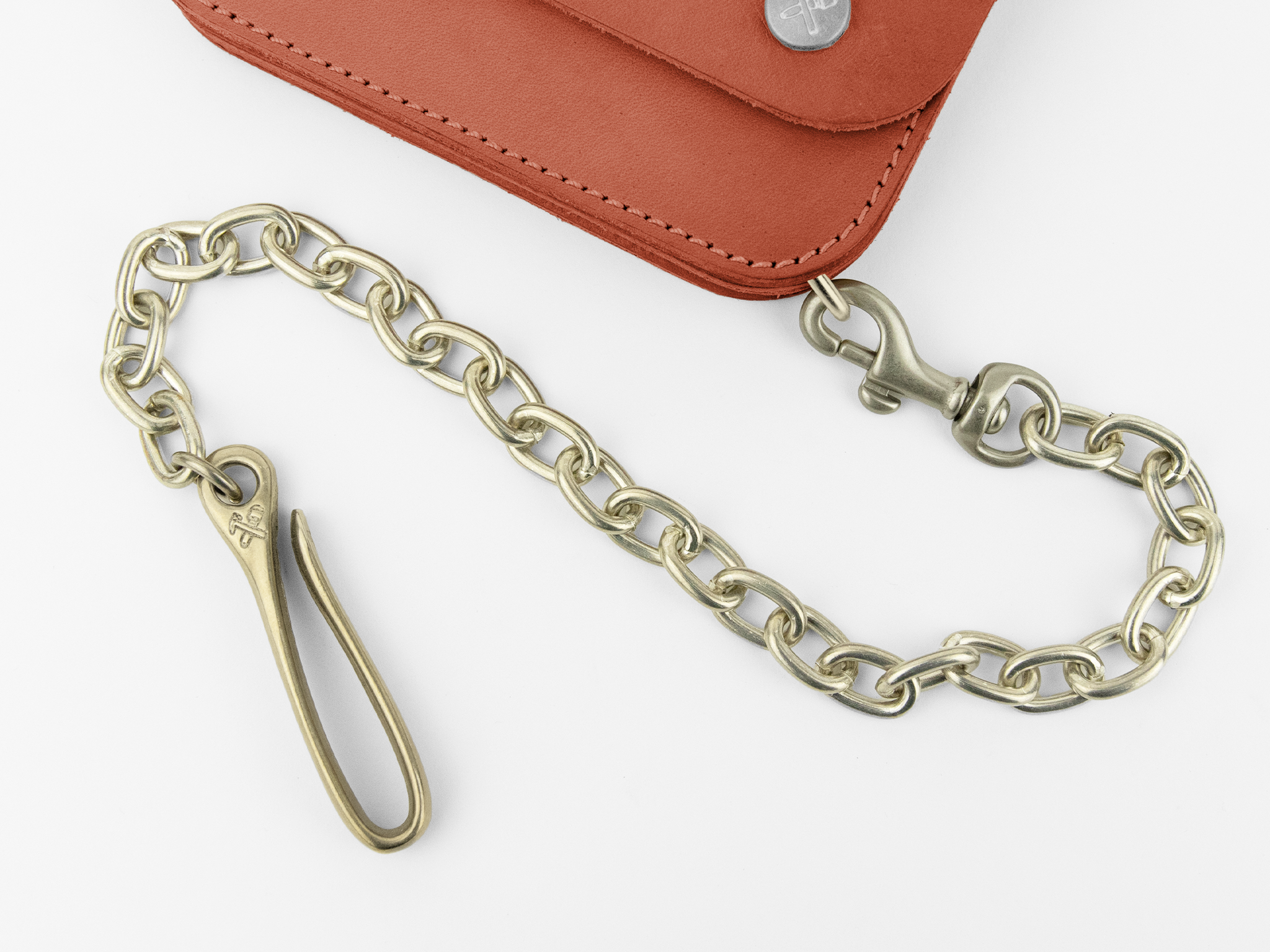 A brass chain with a hook and clasp is attached to a brown leather wallet with visible stitching and metal rivets. The chain is coiled in a loose curve on a white surface.