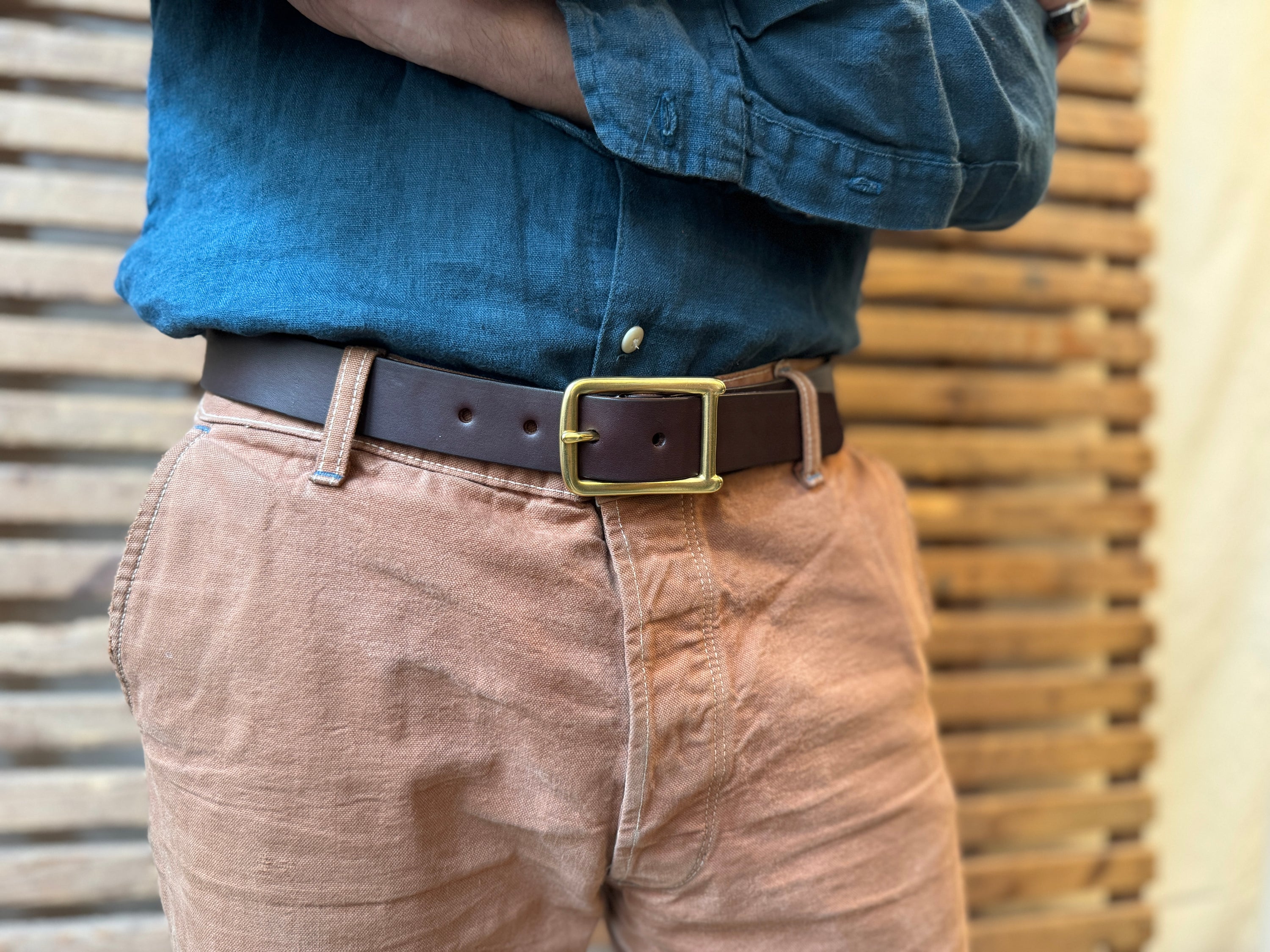 Close-up of a person wearing a blue button-up shirt tucked into light brown pants, secured with a dark brown leather belt featuring a gold buckle. The background consists of horizontal wooden slats.