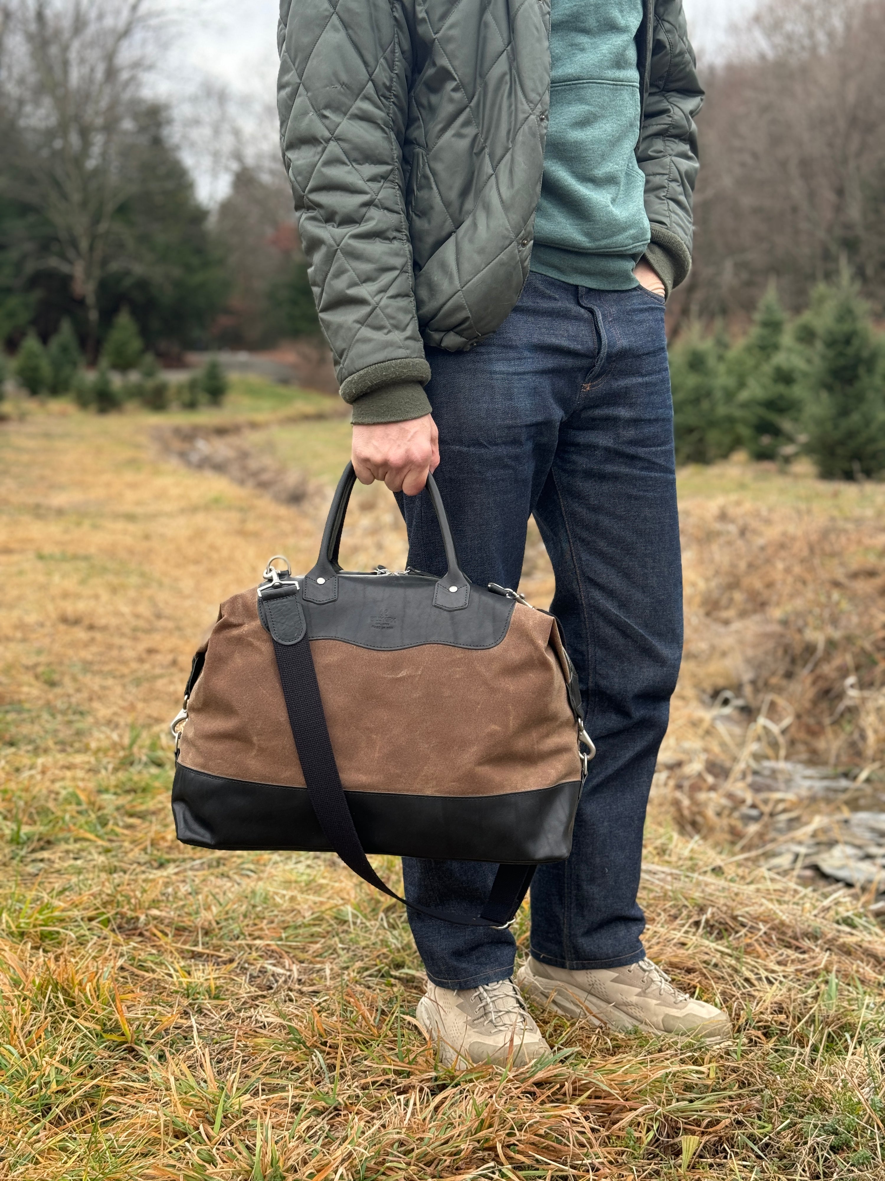 A person standing outdoors on grass, wearing blue jeans, a green quilted jacket, tan shoes, and holding a large brown and black duffel bag with a black strap. Pine trees are visible in the background.