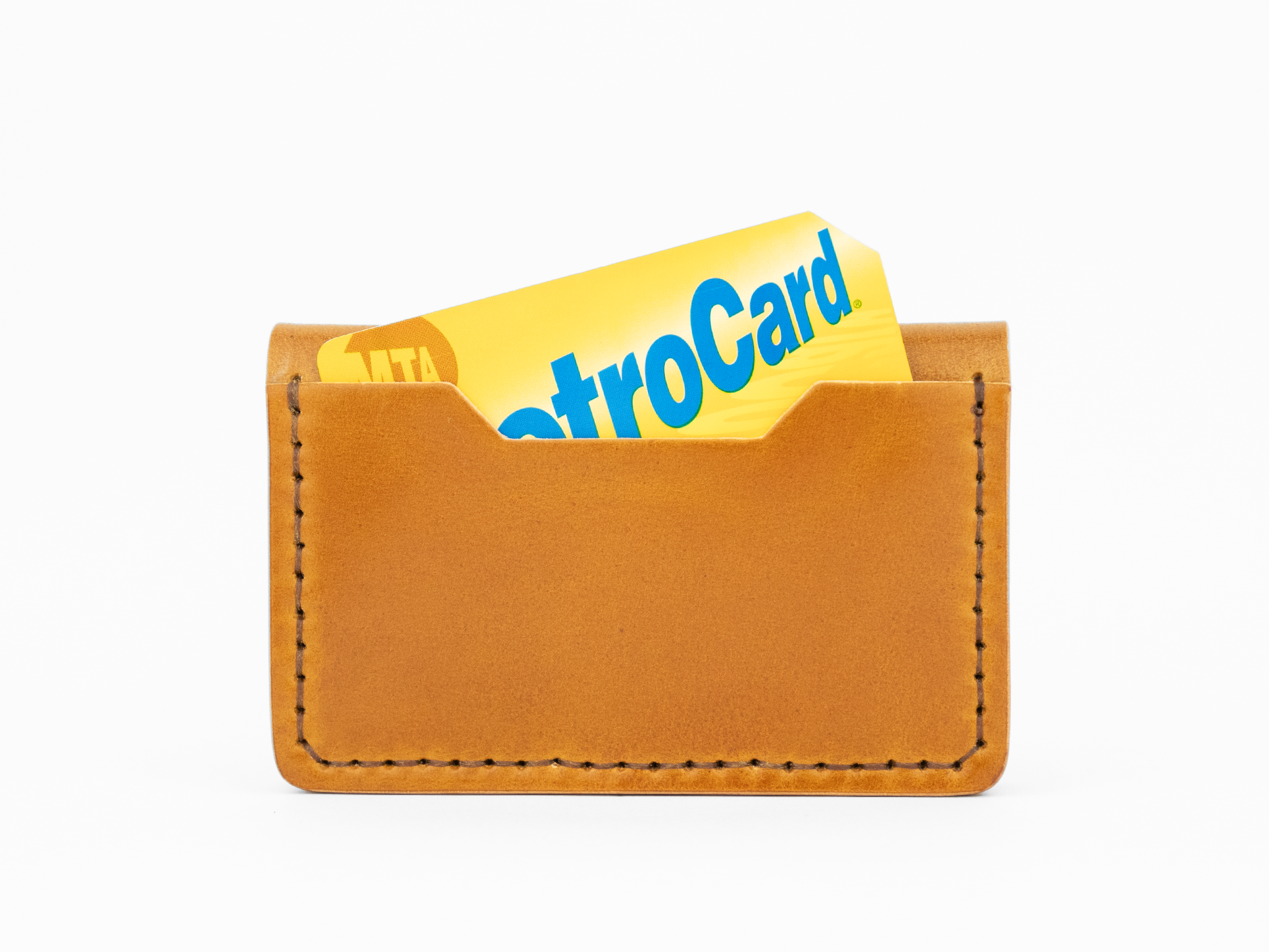 A tan cordovan leather card holder with visible stitching holds a yellow and blue MTA MetroCard, partially sticking out from the top.