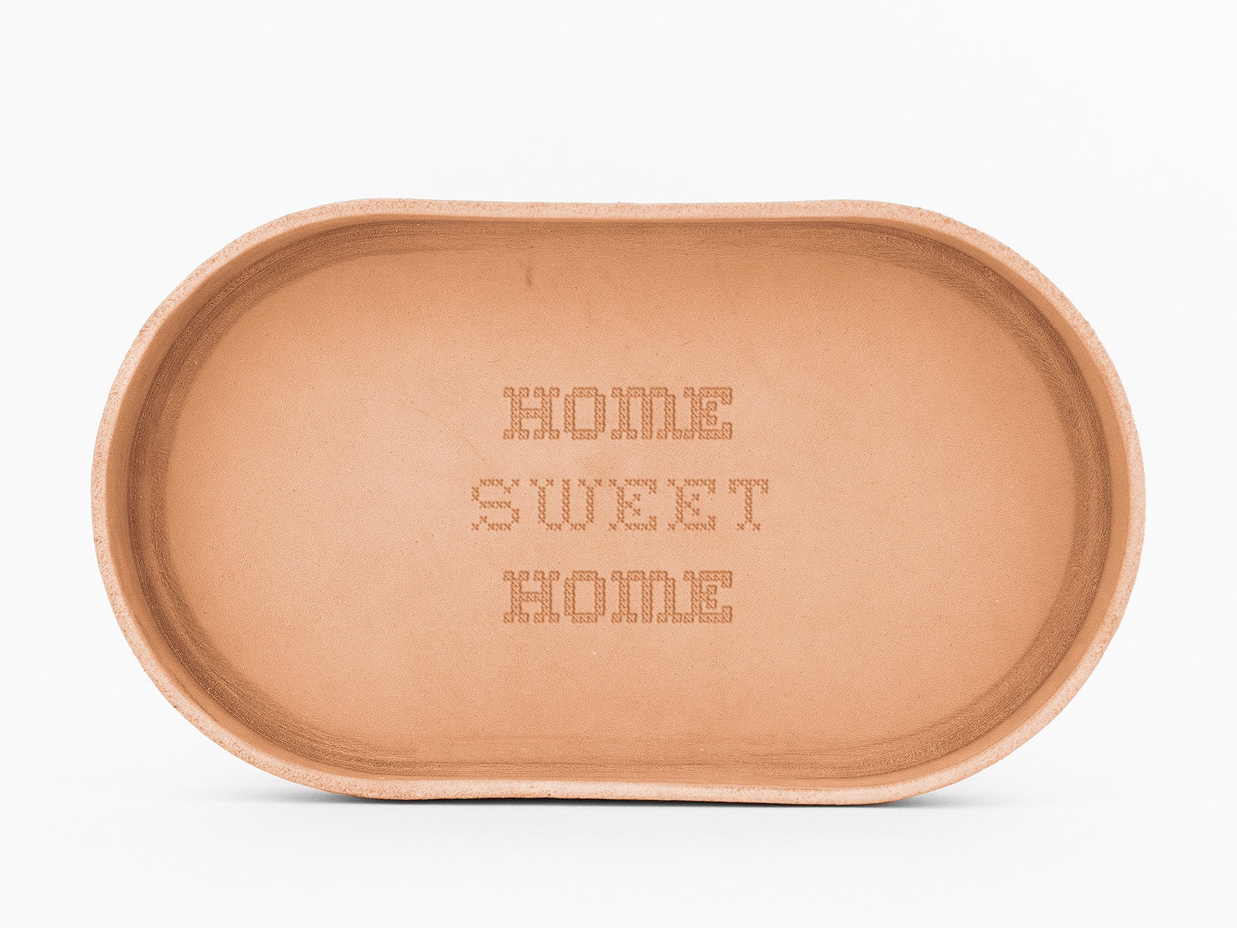 oval-shaped, natural colored leather   tray with the words HOME SWEET HOME engraved in a cross-stitch style in the center. The tray has smooth edges and a simple, rustic appearance.