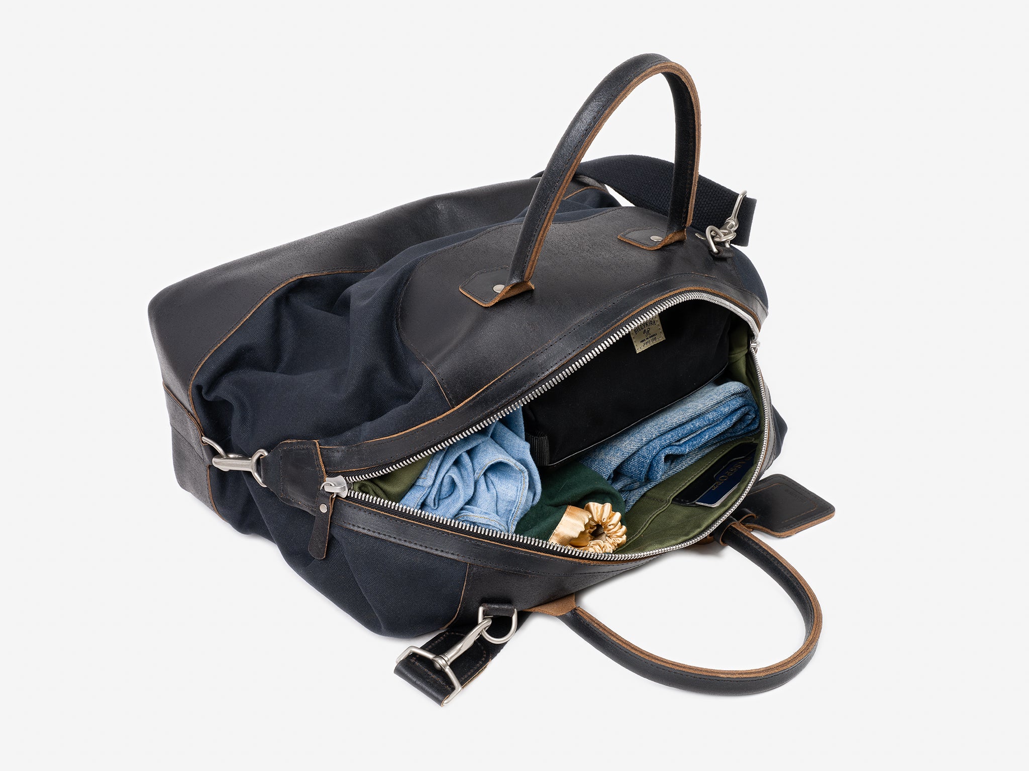 A black leather travel bag with brown trim, partially unzipped, revealing neatly packed clothes and a gold-colored flower brooch inside against a white background.