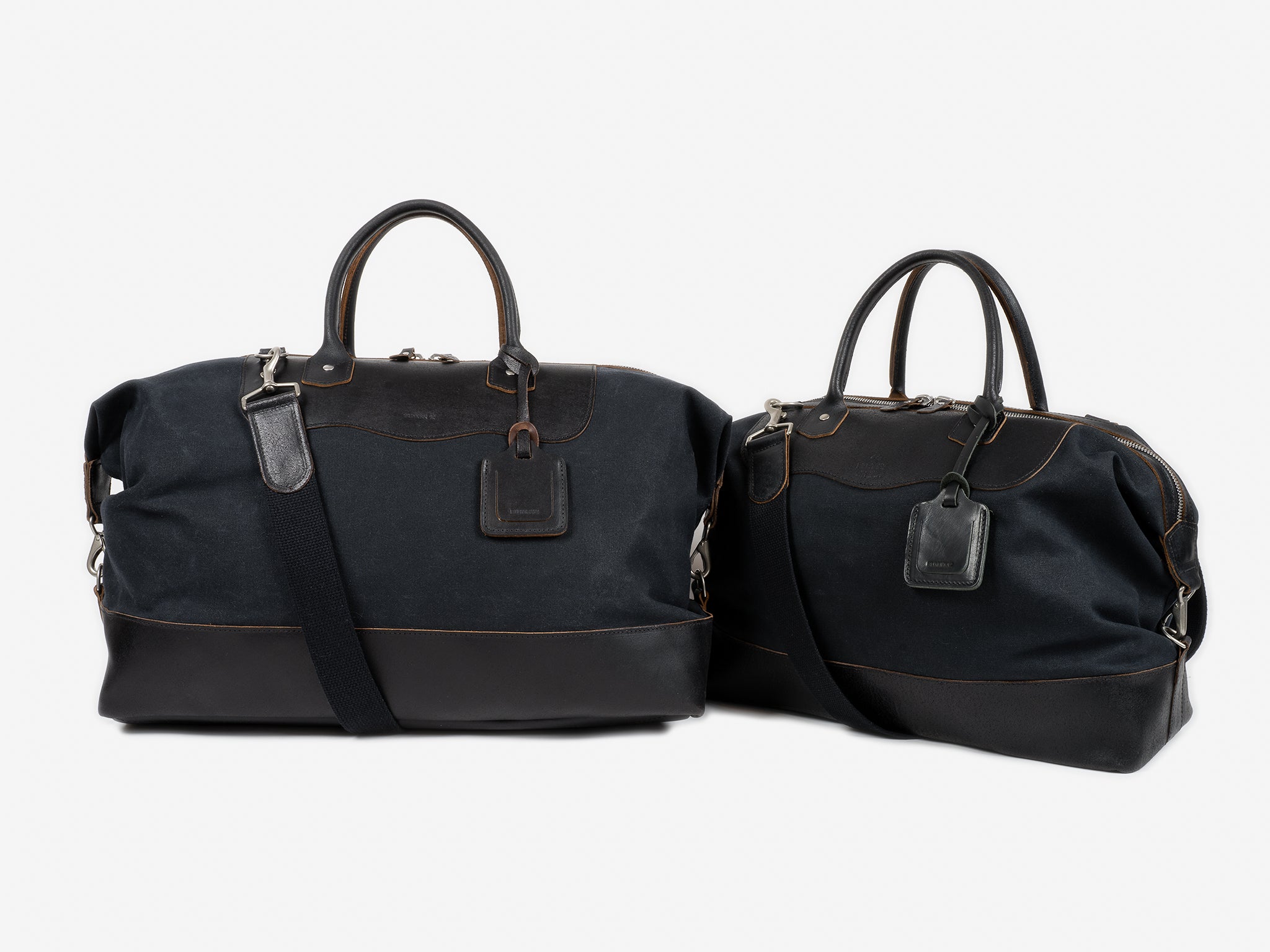 Two black leather and canvas duffel bags with brown stitching, each with handles and an attached luggage tag, are placed side by side on a plain white background.