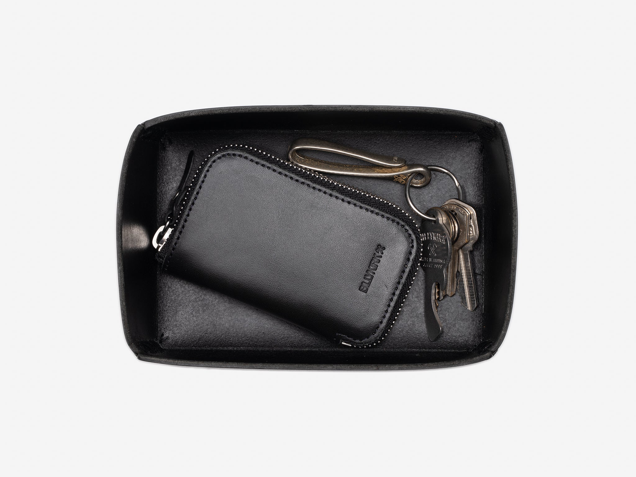 A black leather zippered wallet and a set of metal keys with a brass keychain rest inside a black rectangular open box against a white background.