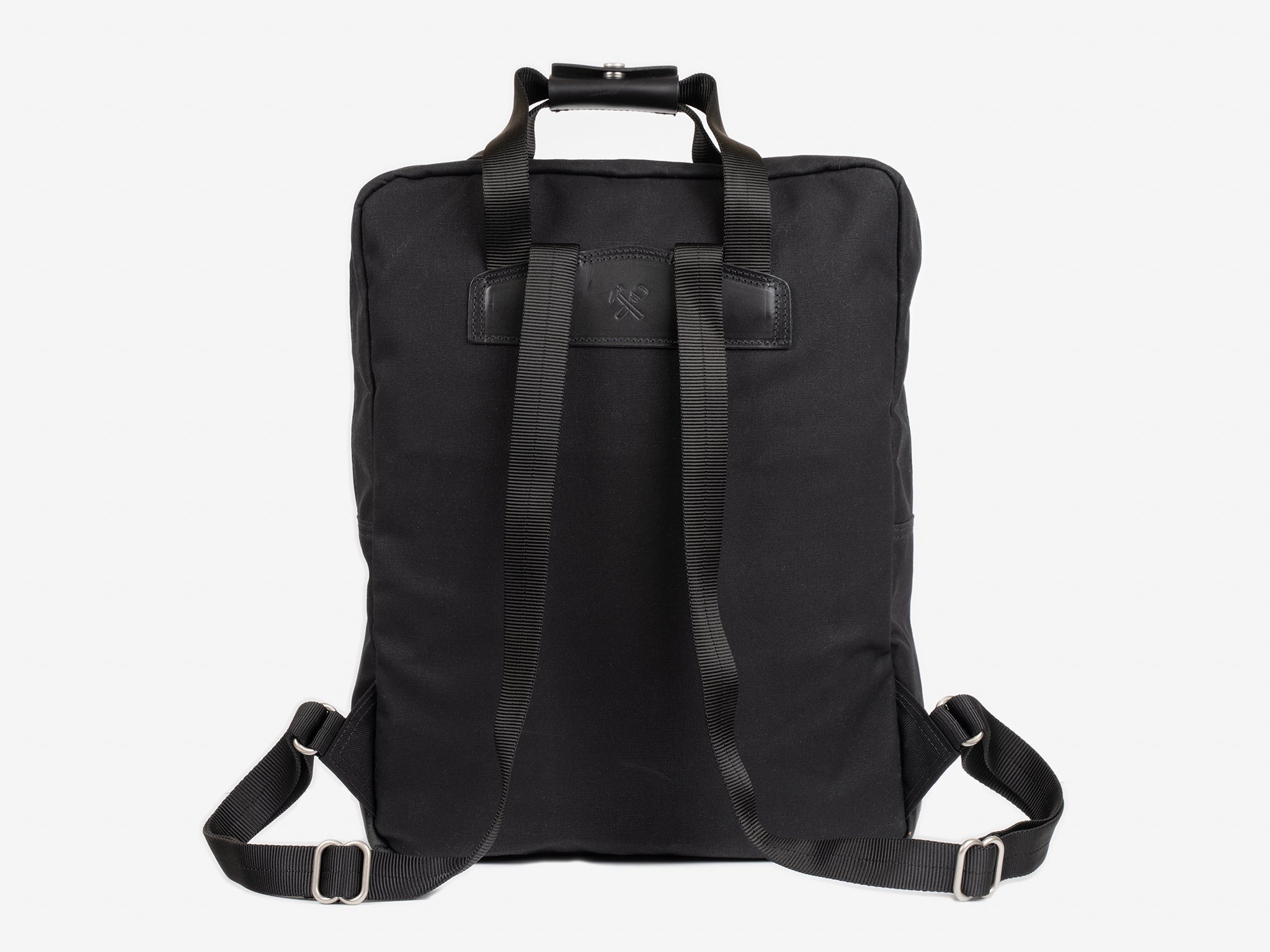 A black waxed canvas backpack shown from the back, featuring adjustable shoulder straps, a top handle, and a rectangular black leather patch near the top center. The background is plain white.