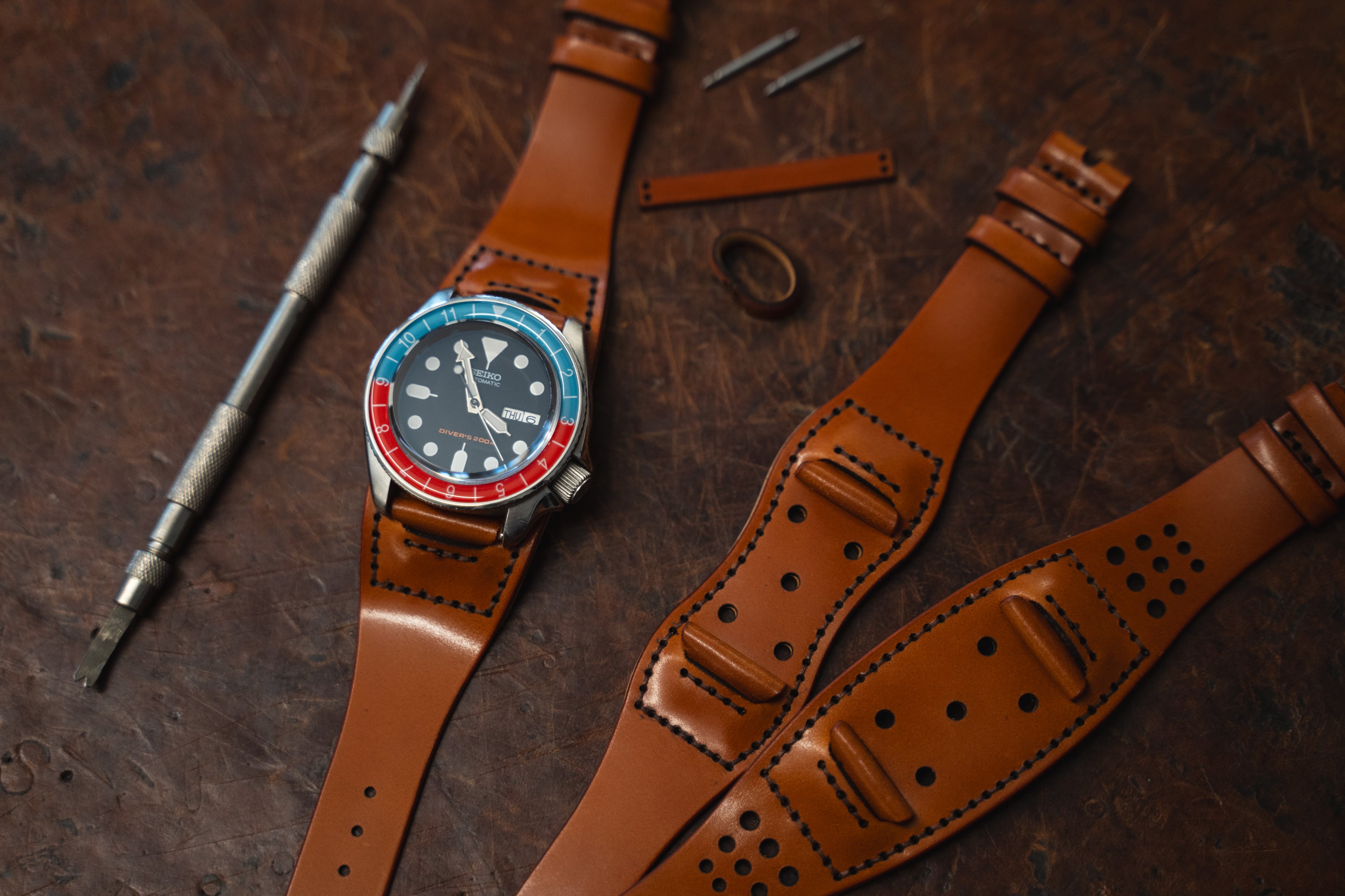 A wristwatch with a blue and red bezel sits on a brown leather strap, surrounded by additional leather straps, a metal tool, watch pins, and a small leather piece on a rustic wooden surface.