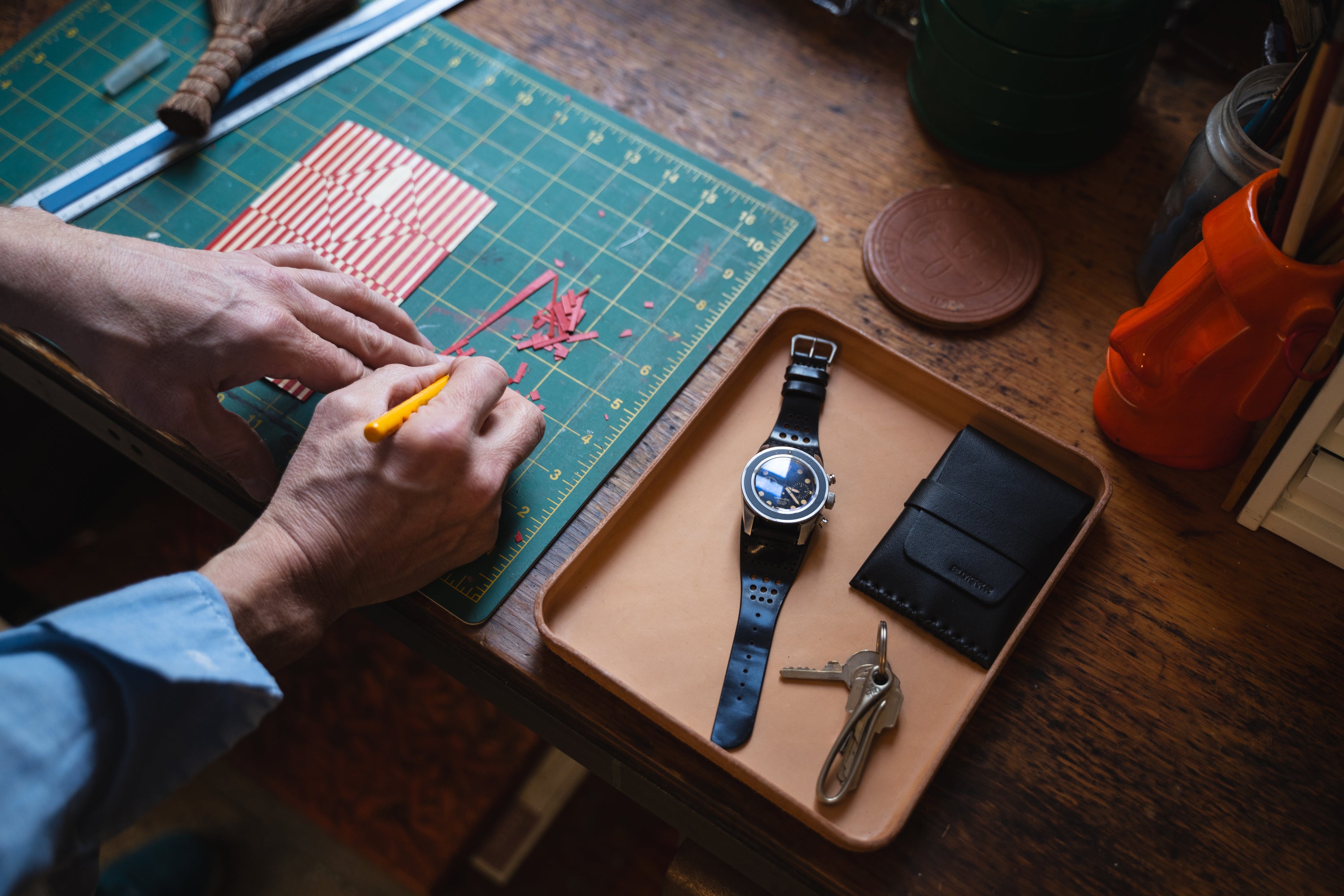 A tray holds a watch with a black cordovan leather strap, a wallet, and keys on a wooden desk. Nearby, a person uses an X-acto knife to cut a red and white striped sheet on a cutting mat.