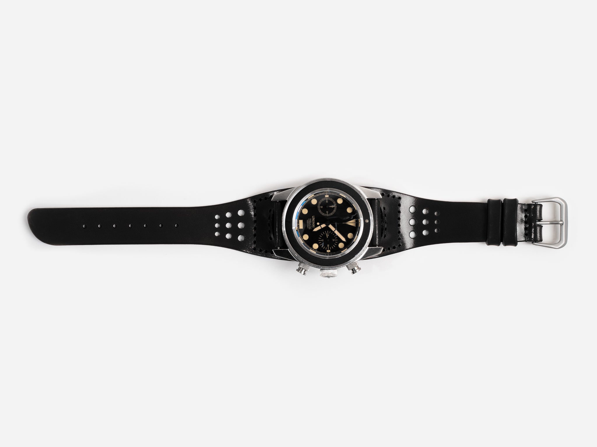 A black cordovan leather watch strap, silver buckle, and a round black dial featuring multiple subdials and gold accents, displayed flat on a white background.