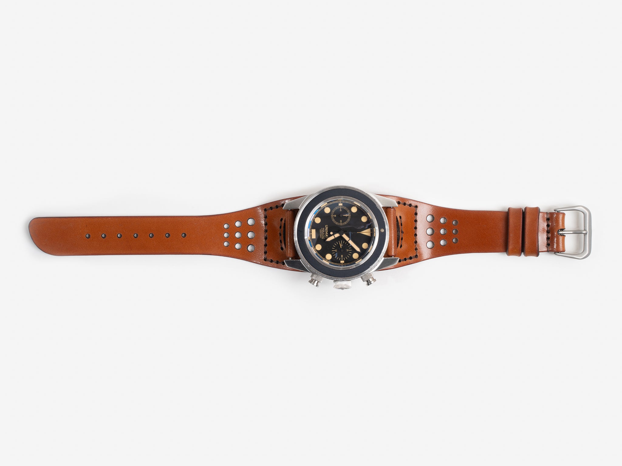 A tan cordovan leather watch strap, silver buckle, and a round black dial featuring multiple subdials and gold accents, displayed flat on a white background.