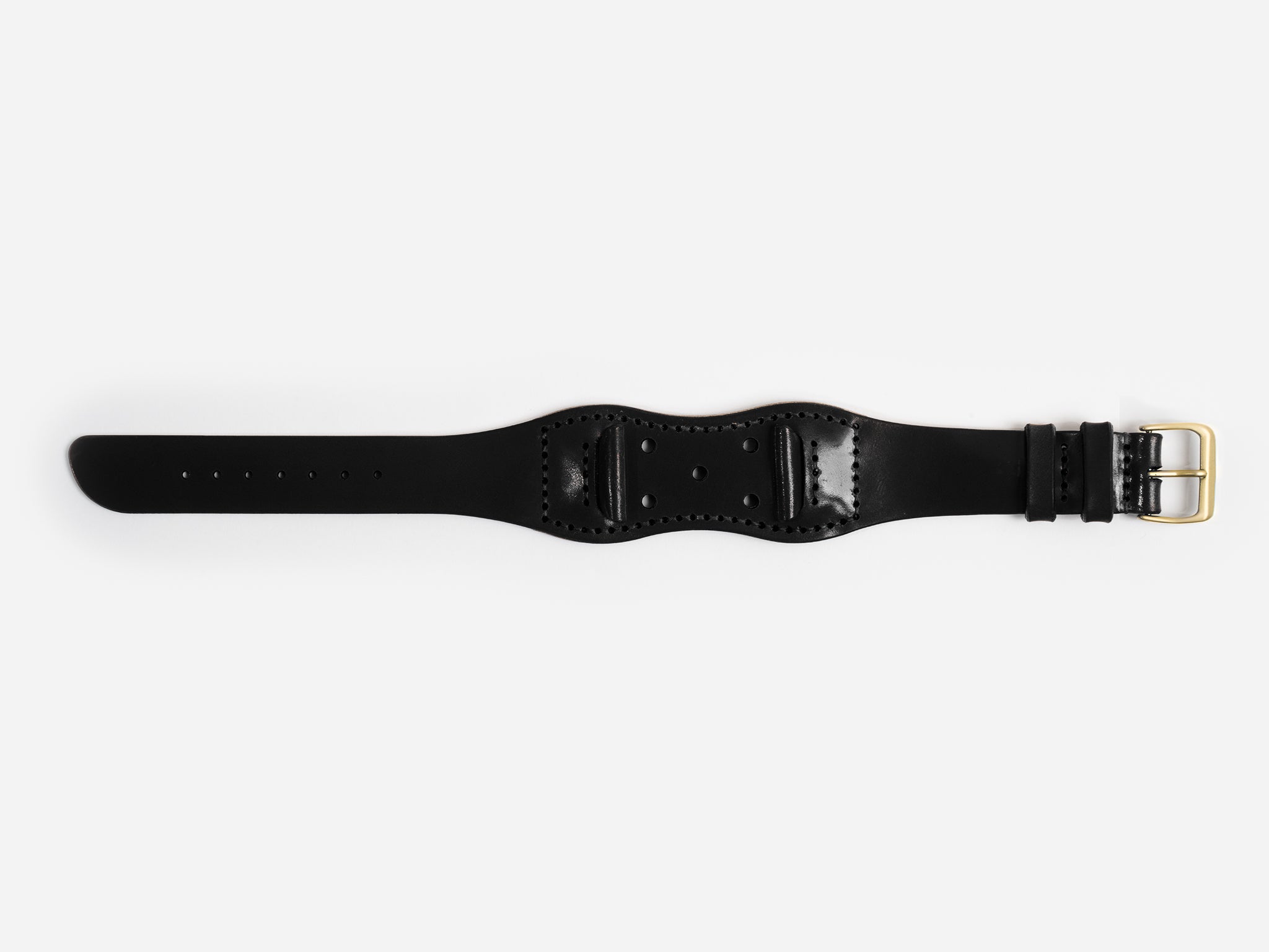 A black cordovan leather watch strap with a brass buckle lies flat on a white background; it has stitched edges and an hourglass shape.