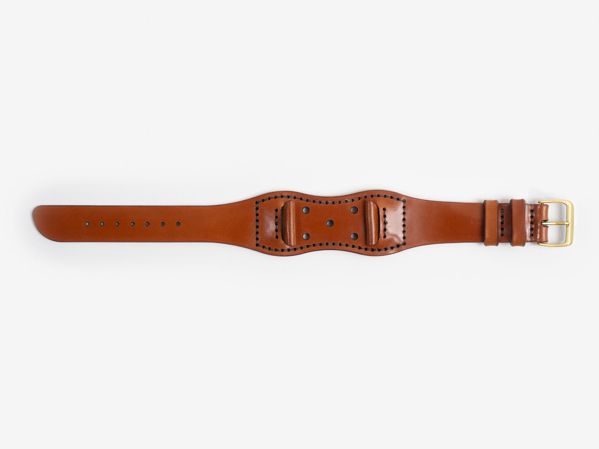A tan cordovan leather watch strap with a brass buckle lies flat on a white background; it has stitched edges and an hourglass shape.