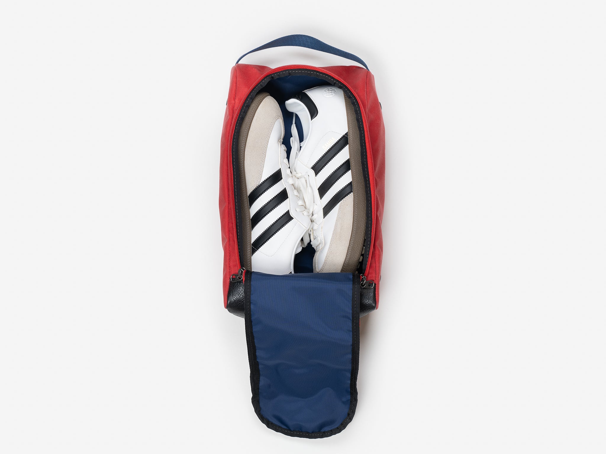 A pair of white sneakers with black stripes placed inside a red shoe bag with a navy blue interior and the top flap partially unzipped.