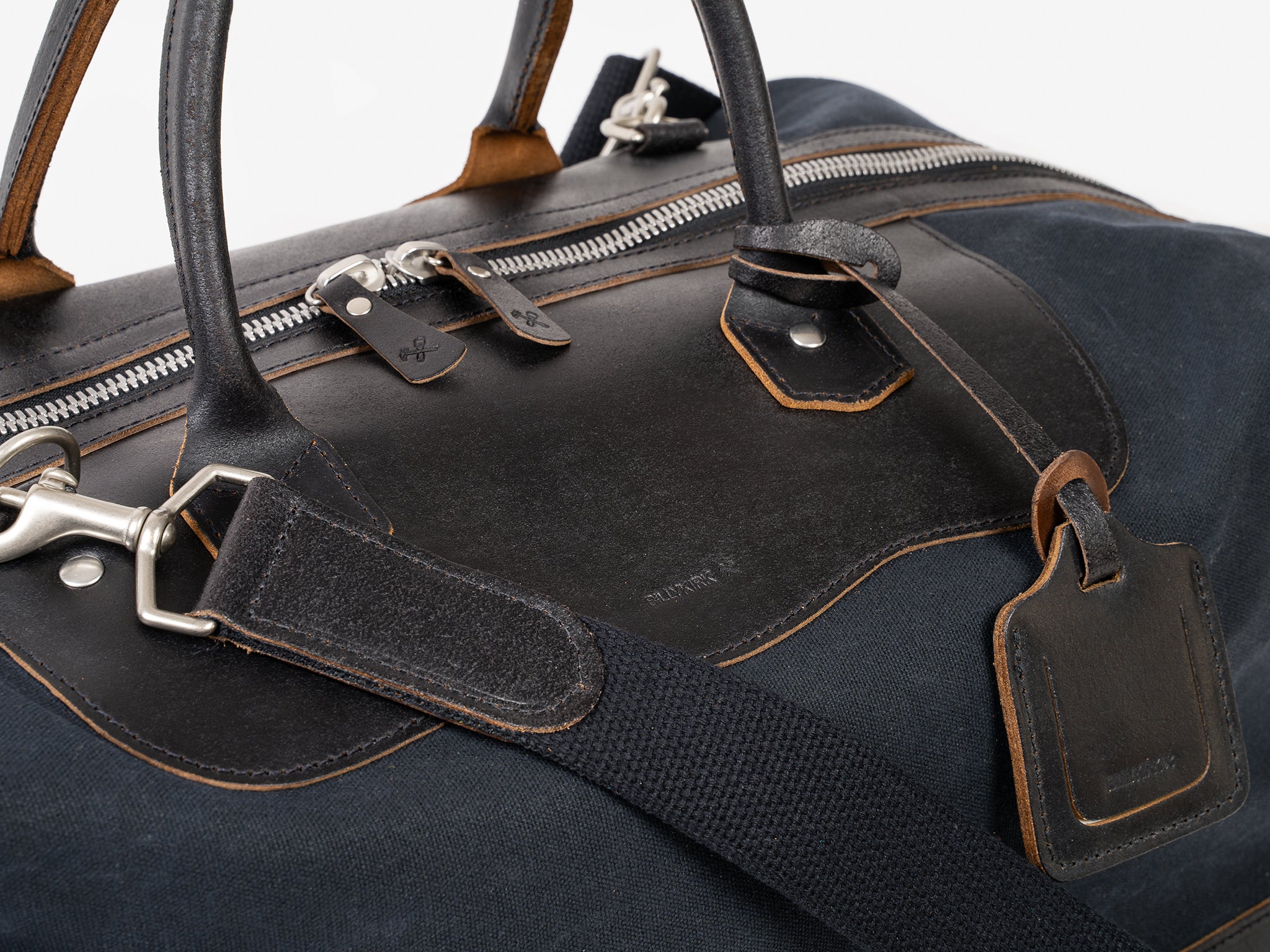 A close-up view of a dark blue and black leather travel duffel bag, featuring sturdy handles, a shoulder strap, metal zipper, and a luggage tag. The bag has visible stitching and silver-tone hardware.