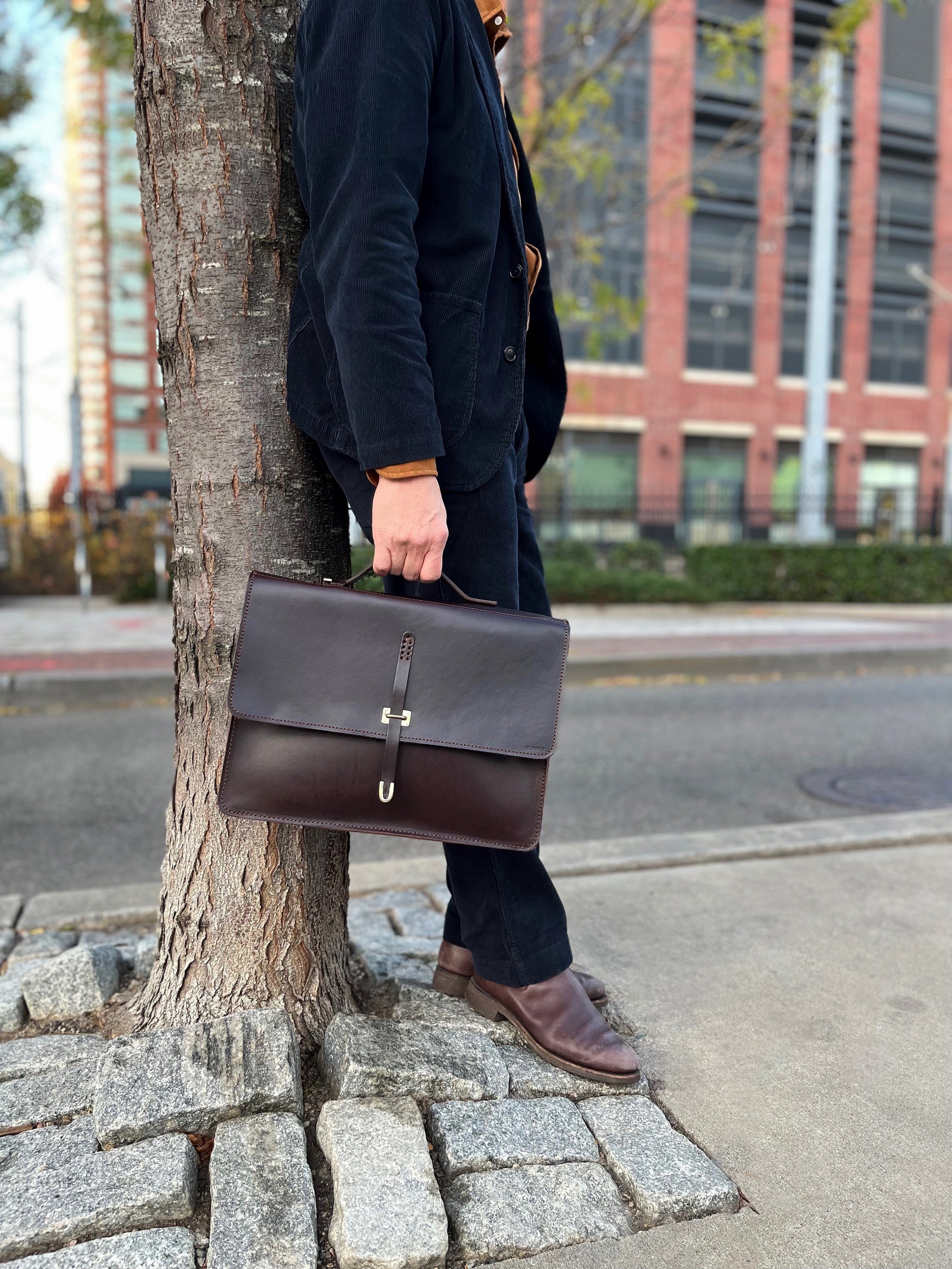 A person in dark clothing leans against a tree on a city sidewalk, holding a dark brown leather briefcase. Only the lower half of their body is visible. A red brick building and street are in the background.