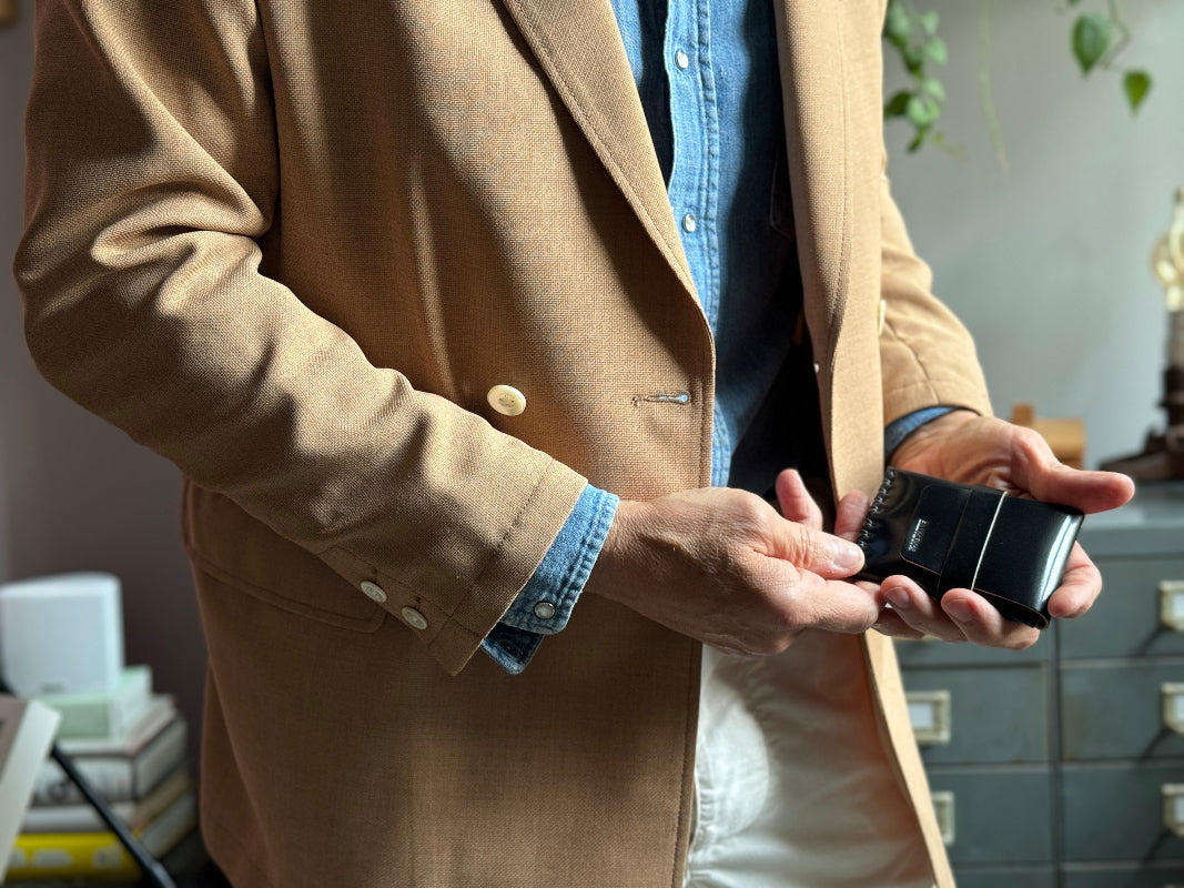 A person in a tan blazer, blue shirt, and white pants holds a black vintage camera in their hands, standing indoors near a stack of books and a set of drawers with plants in the background.