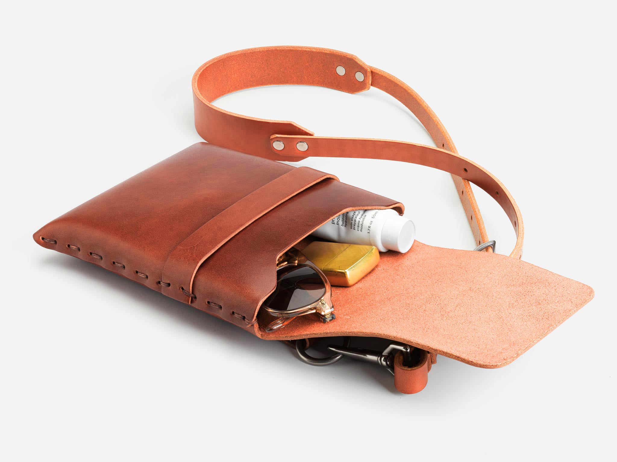 A brown leather crossbody bag with the flap open, showing eyeglasses, a bar of soap, and a small tube inside. The bag has a thin shoulder strap and is displayed on a white background.