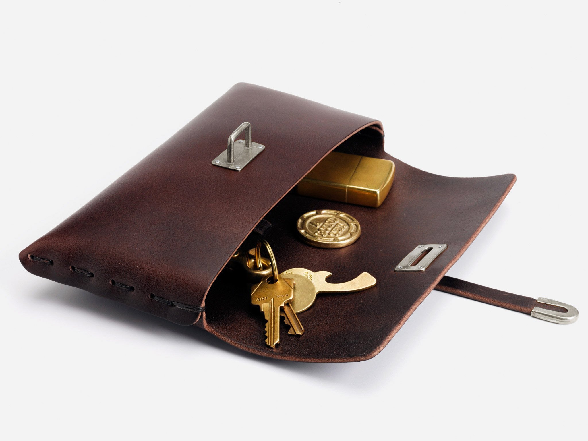 A brown leather pouch with a metal clasp, lying open on a white surface, revealing keys, a round coin, and a rectangular brass box inside.