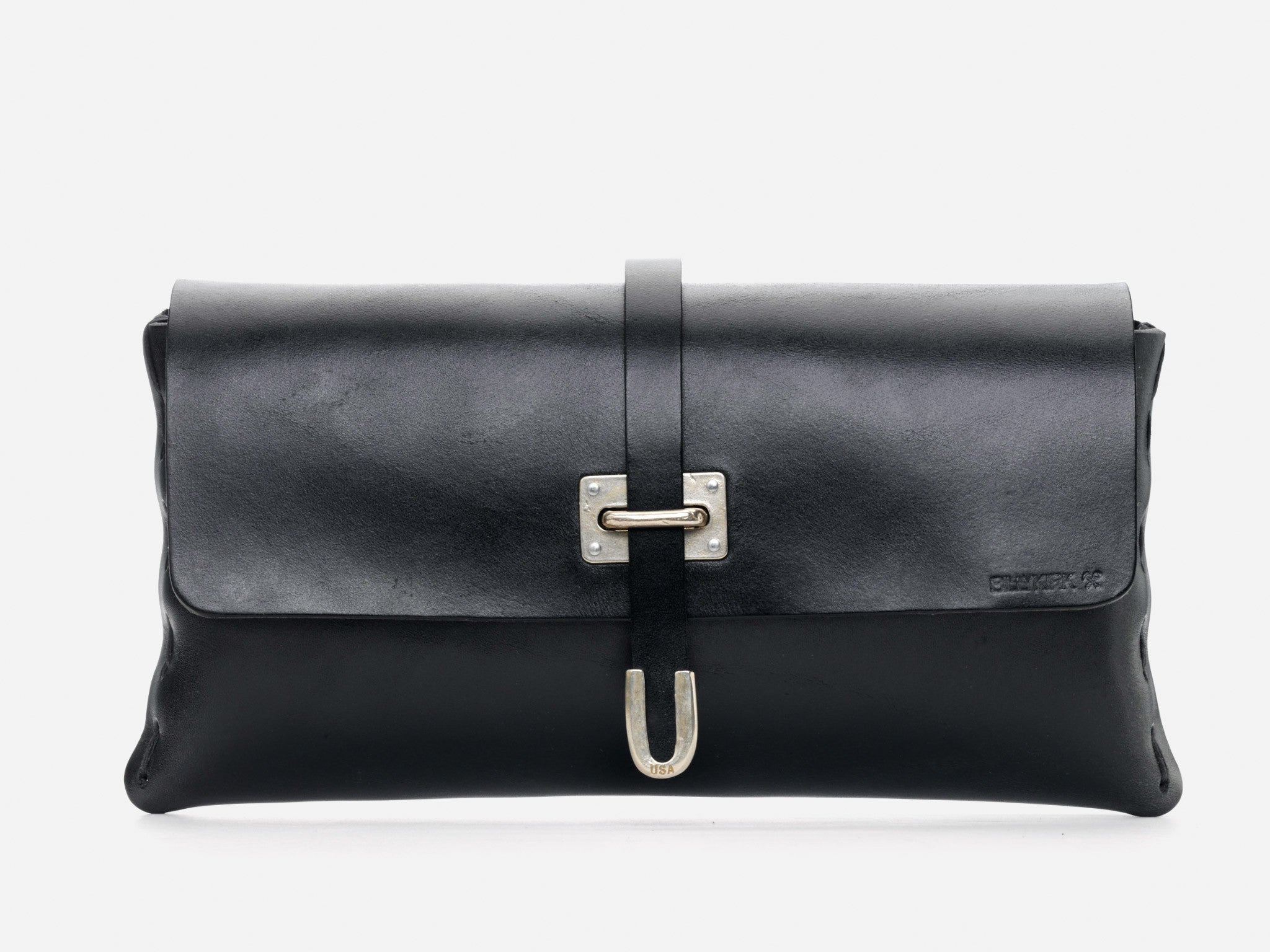 A black leather clutch bag with a fold-over flap, secured by a strap and a silver buckle. The surface is smooth, and the brand name is embossed on the lower right corner. The background is white.