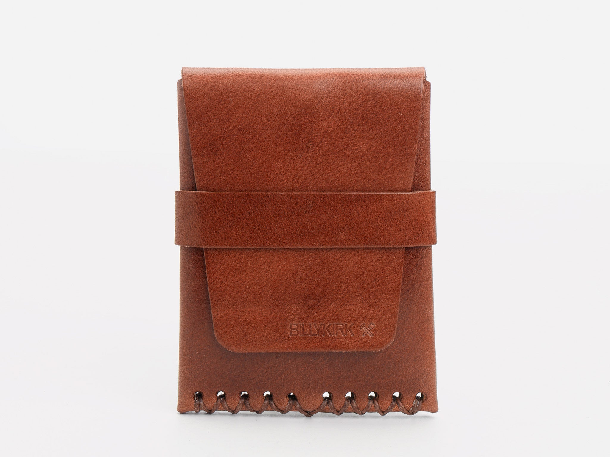 A tan leather card case with a flap closure, secured by a horizontal strap and stitching along the bottom edge. The card case has a minimalist design and subtle embossed branding on the lower front.