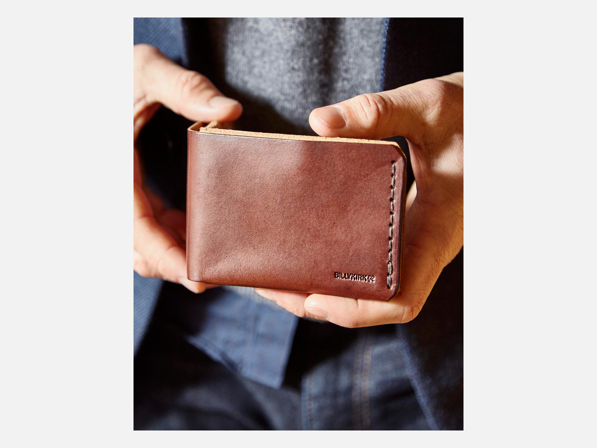 A person wearing a blue jacket and grey shirt holds a brown leather wallet with neat stitching and the word BULLSTRAP embossed in the corner.