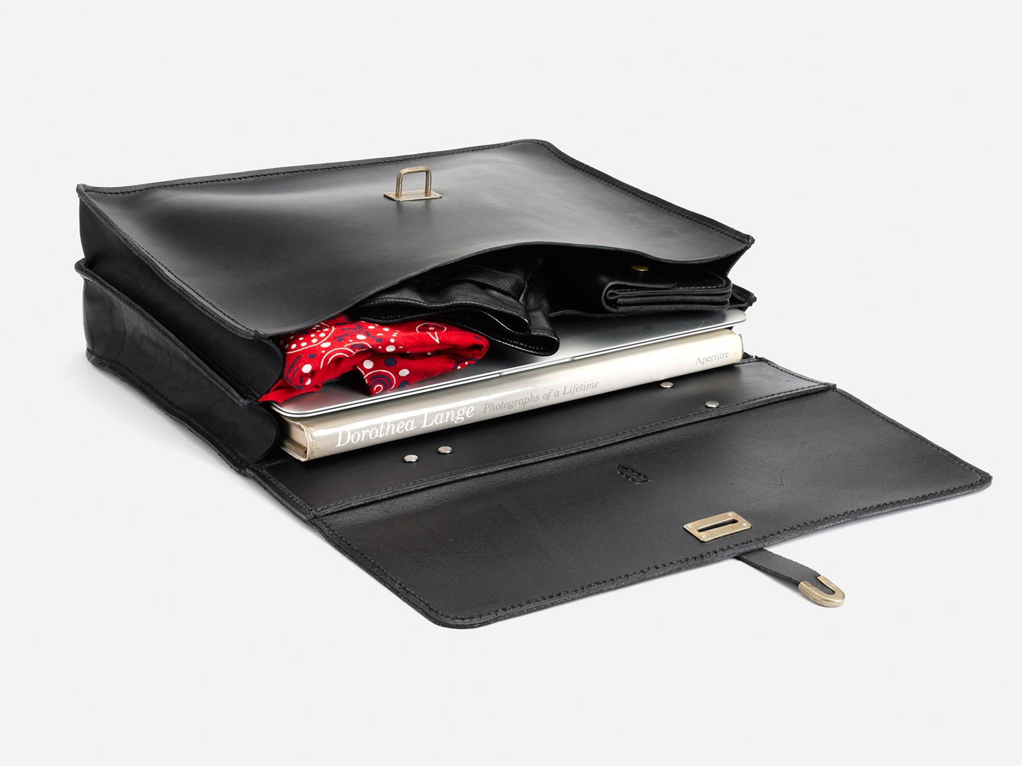 An open black leather satchel with a red bandana, a rolled-up magazine, and a hardcover book titled Dorothea Lange neatly packed inside, displayed on a white background.