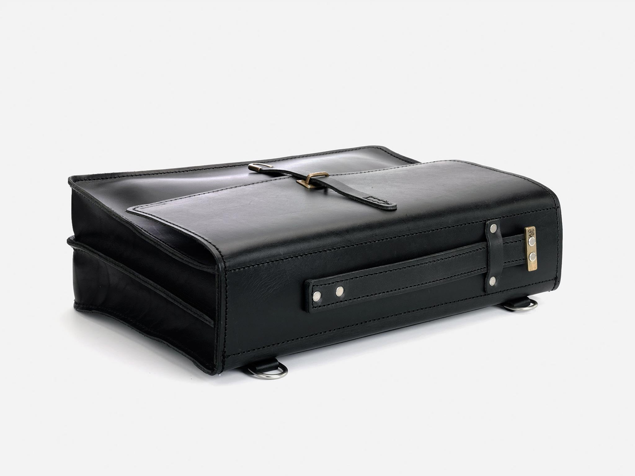 A black leather briefcase lies flat on a white surface. It features a top buckle strap, side handle, metal feet, and a combination lock on one end.