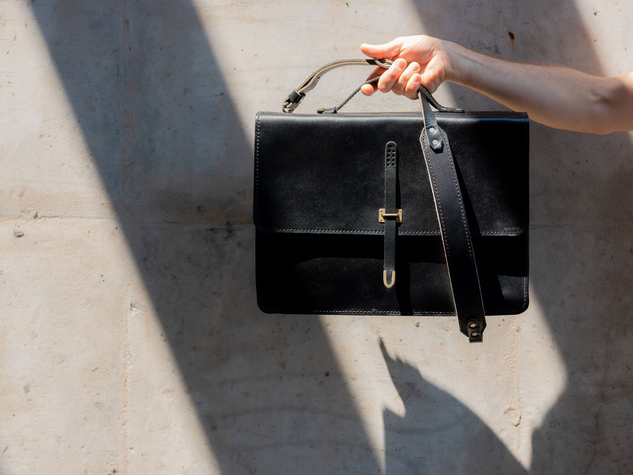 A person’s hand holds up a black leather briefcase with a buckle, with sunlight casting strong shadows on a light-colored concrete wall in the background.