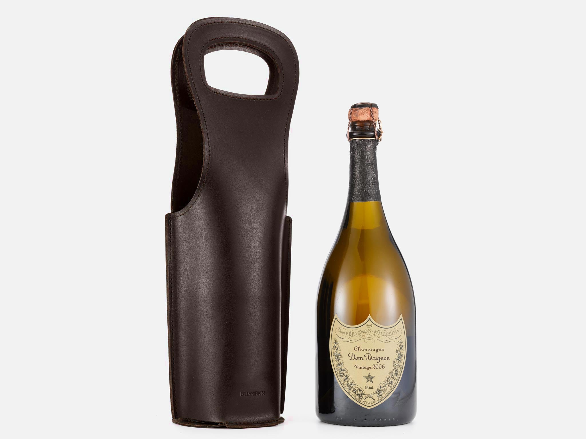 A bottle of Dom Pérignon vintage 2008 champagne stands next to a tall, dark brown leather wine carrier with a handle, both set against a plain white background.