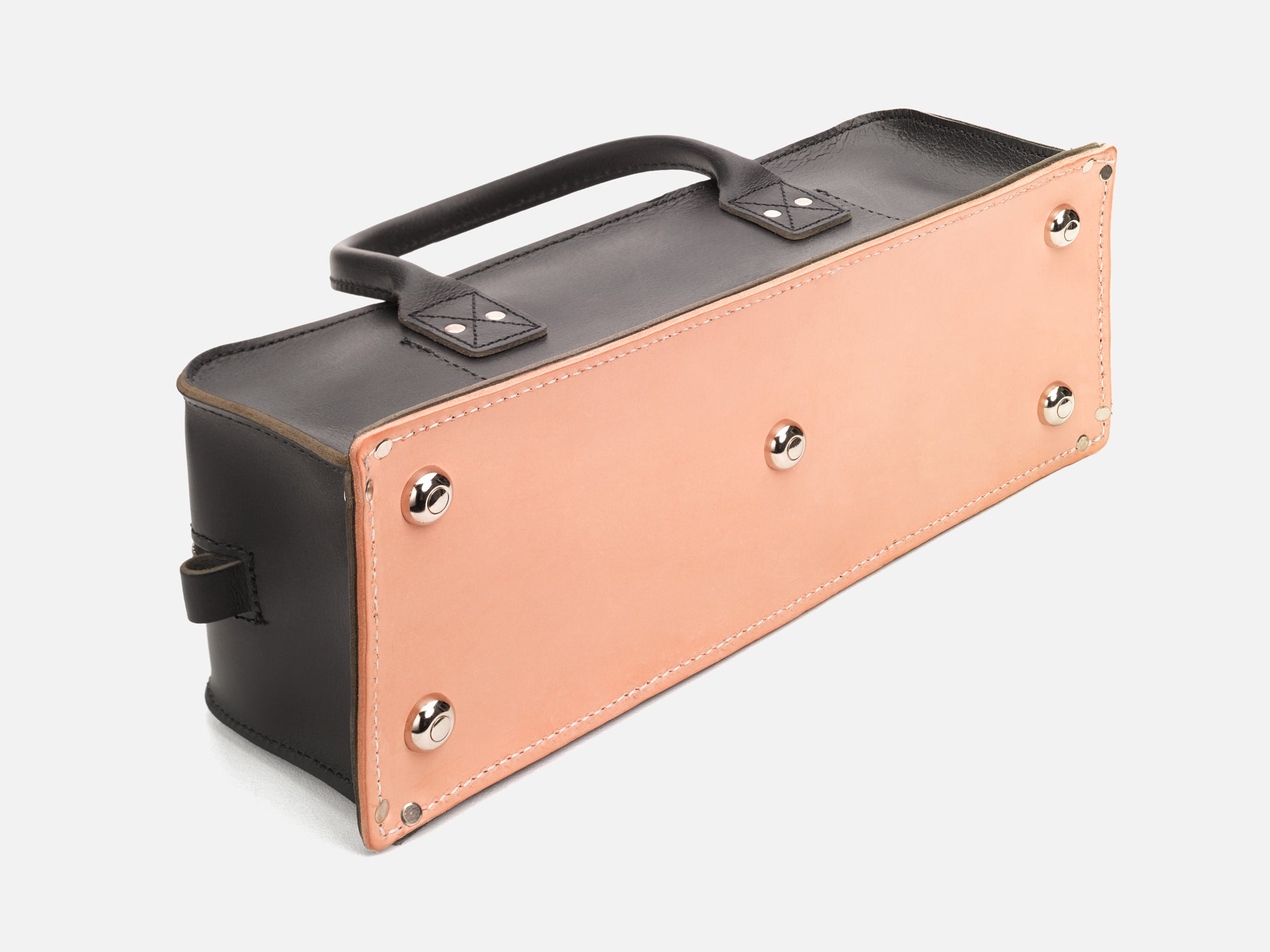 A bottom view of a black leather tool bag with rivet reinforced corners showing the natural veg tan bottom and silver feet set in a white background
