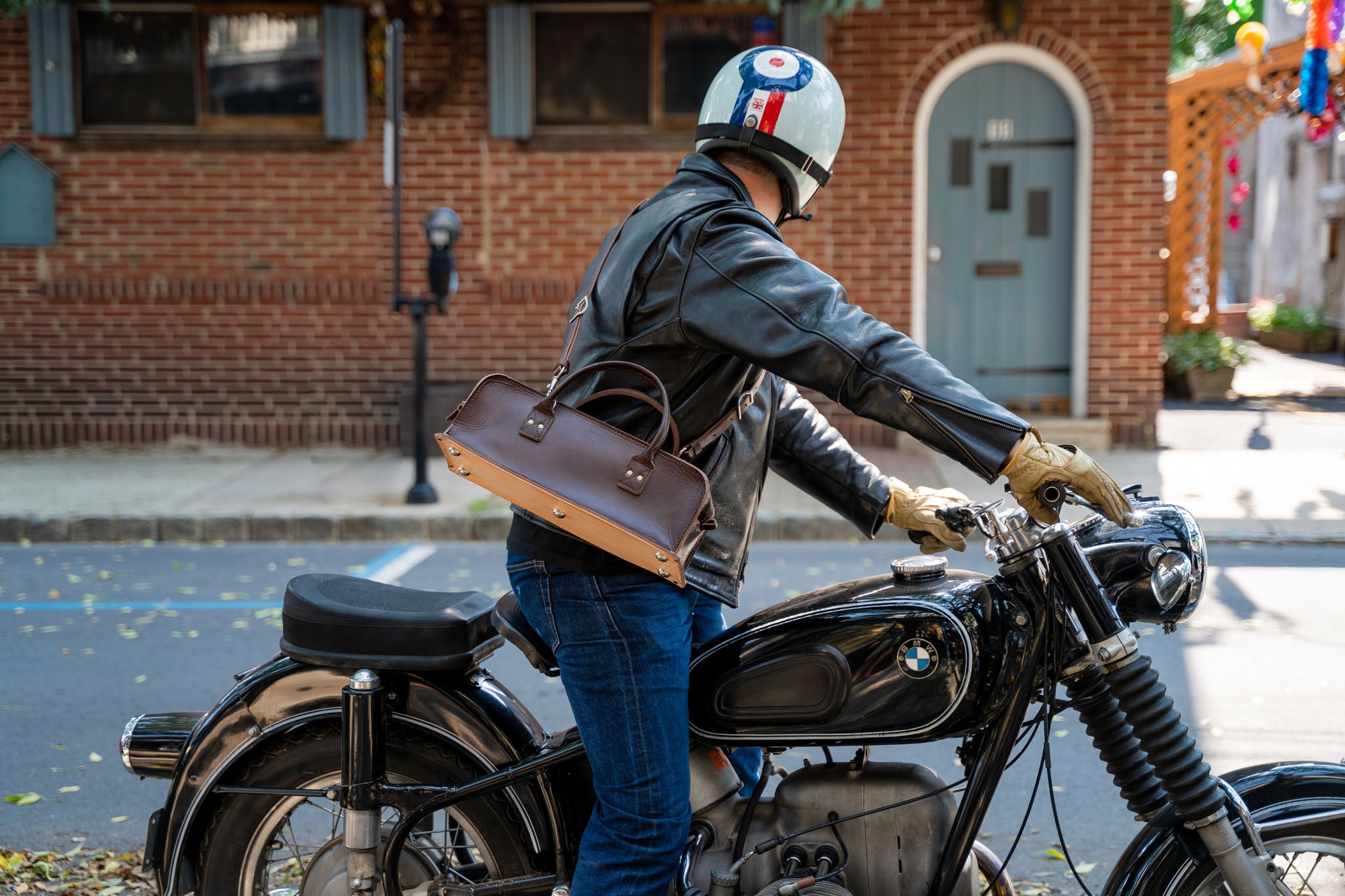 A person wearing a leather jacket and helmet sits on a vintage BMW motorcycle, carrying a brown leather bag, on a city street with brick buildings in the background.