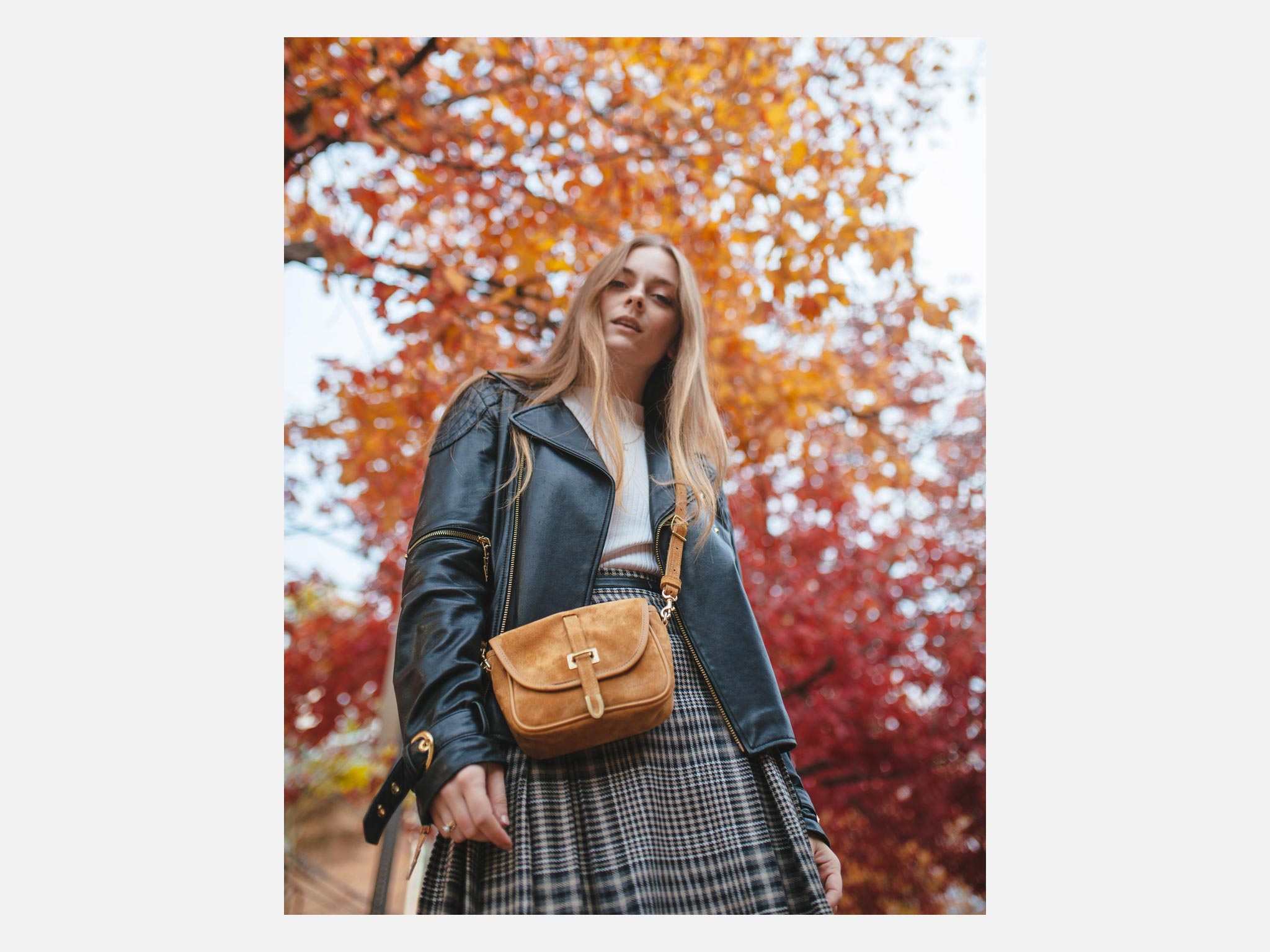 A woman in a black leather jacket and plaid skirt stands outdoors with a yellow crossbody bag, surrounded by vibrant autumn trees with red and orange leaves. She gazes confidently at the camera.